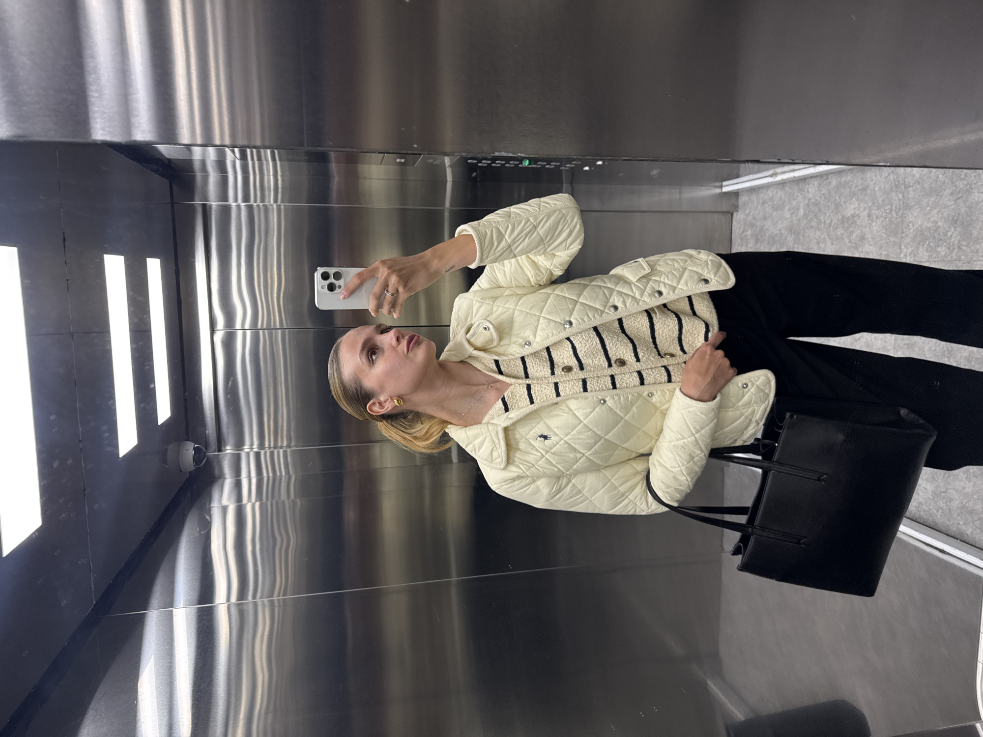 The image depicts a woman in an elevator. The woman is taking a selfie with her smartphone. She's wearing a cream-colored quilted jacket over a striped sweater, black pants, and a black handbag. The elevator has stainless steel walls and a security camera. The floor of the elevator appears to be a light gray. The location is unknown.
