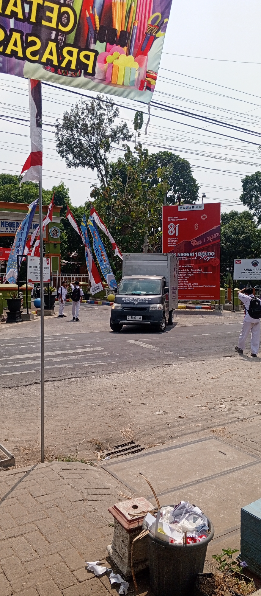 In Maospati, Indonesia, a dark gray delivery truck with license plate D 8995 WE is stopped at a crosswalk in front of a school building. The truck has text on its side that reads "SATUAN PELAYANAN PEMENUHAN GIZI." Two male students in white uniforms are standing on the sidewalk to the left of the truck, one facing forward and the other looking to the right. A large red banner with text in Indonesian is visible on the right side of the image, and several smaller Indonesian flags are displayed on poles in front of the school entrance. The sky is clear and blue, and the ground is paved with asphalt and concrete. A trash can filled with papers is located on the right side of the foreground.