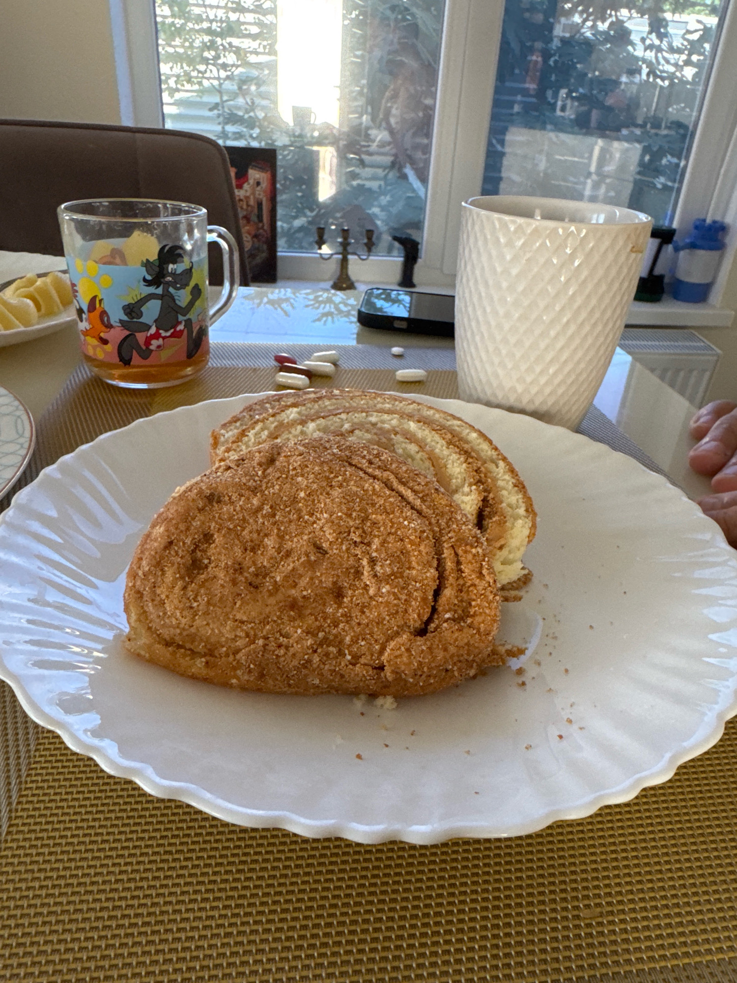 A close-up, overhead shot shows a white, scalloped plate with a swirled pastry covered in a brown, crumbly topping. The pastry is sliced, revealing layers of dough and a lighter filling. In the background, to the left, a clear glass mug filled with a beverage and adorned with cartoon characters sits on a textured placemat. Behind the mug, a dark brown chair is visible. To the right of the mug, a white ceramic cup with a diamond pattern is positioned on the placemat. Between the mug and the ceramic cup, several pills, some white and some brown, are scattered on the placemat. A smartphone lies face down near the pills. The background features a window with sunlight streaming through, revealing trees and greenery outside. The scene appears to be indoors, on a table covered with a woven placemat, likely in a home in the city of Uyutnoye, Ukraine.
