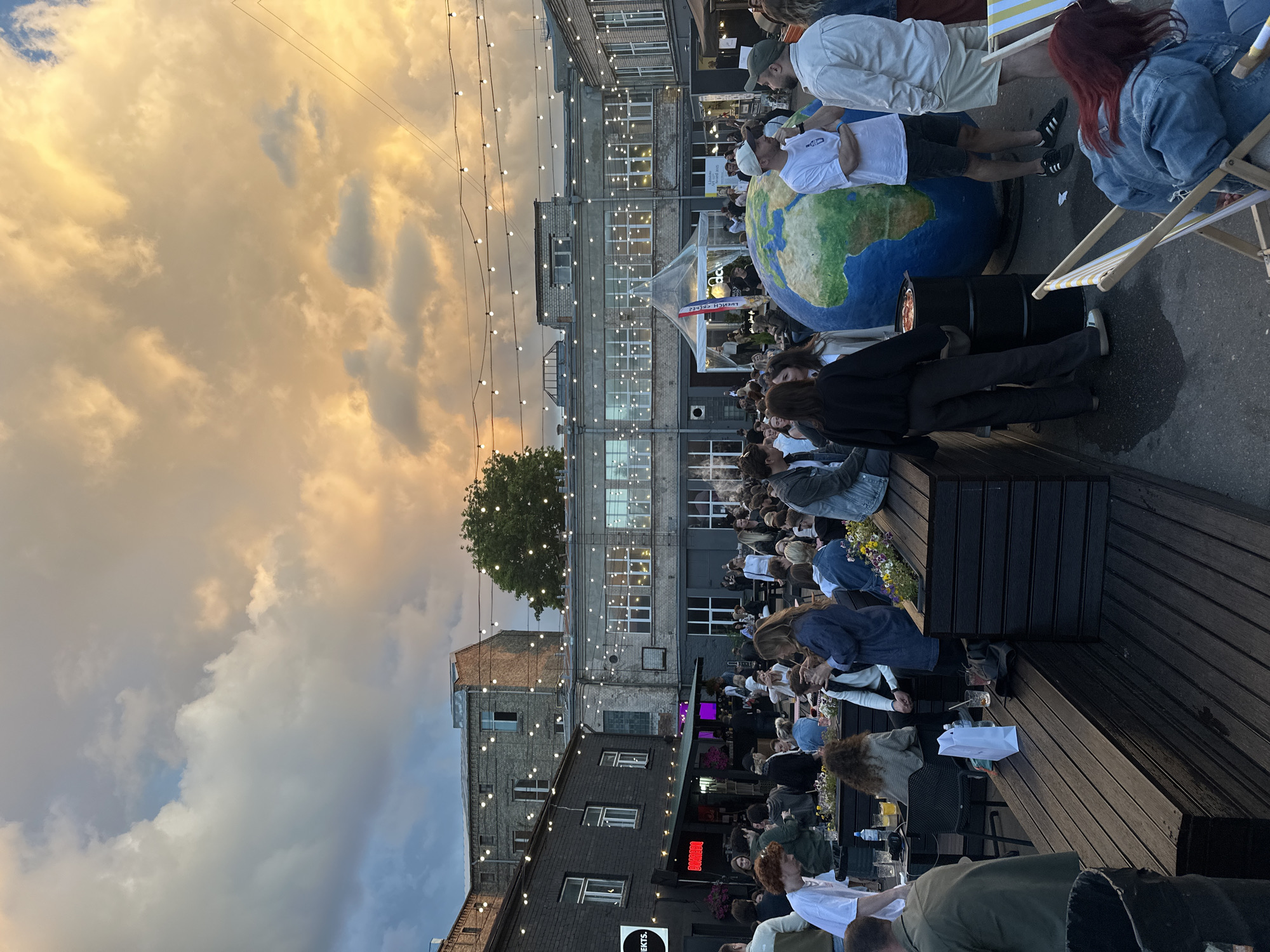 This detailed description pertains to an outdoor social event occurring in the late afternoon or early evening at Tallinas Kvartāls (Tallinn Yard Quarter) in Riga, Latvia. The scene depicts a large, open-air courtyard surrounded by multi-story, industrial-style brick and concrete buildings. Some buildings are dark grey or black, while others are lighter grey or reddish-brown brick. Numerous large windows are visible on the building facades. String lights with bare bulbs are suspended in multiple rows across the sky, crisscrossing the courtyard, and are also strung along awnings and building edges. The sky above is partially overcast with dramatic clouds; the left side features blue-grey clouds, transitioning to warm, golden-orange and yellow hues on the right, suggesting either sunset or intense light from the setting sun. A significant crowd of people, predominantly young adults, are gathered throughout the courtyard. They are casually dressed in a variety of attire, including t-shirts, hoodies, jeans, and light jackets. Many individuals are standing in small groups, engaging in conversations and socializing. Others are seated at dark wooden tables and on built-in wooden benches that define various seating areas. Several people are holding glasses containing drinks, some appearing to be alcoholic beverages or cocktails. In the mid-ground, a prominent, large, spherical object resembling a globe of the Earth is positioned. It is predominantly blue for oceans and green/brown for continents. Near the globe, two men are conversing; one wears a white t-shirt, dark shorts, and a white baseball cap, while the other wears a light-colored shirt and shorts, and a dark baseball cap. Close by, a black barrel topped with a small red container and possibly a grill is visible. Various temporary structures, likely food and drink stalls or pop-up shops, are visible in the background. One notable structure is a white tent with a clear plastic top, featuring a red and blue flag attached. A darker-colored stall, potentially a food truck, is also present further back. A sign partially reading "EKTS." is visible on a dark building to the left, and another sign reading "BOMBEL" is under an awning. The ground is paved, appearing somewhat wet or reflective in certain areas. To the lower left, a black trash receptacle is visible. In the lower right foreground, a person with red hair is relaxing in a yellow and white striped deck chair. Small planters containing flowers are integrated into some of the wooden seating structures, adding