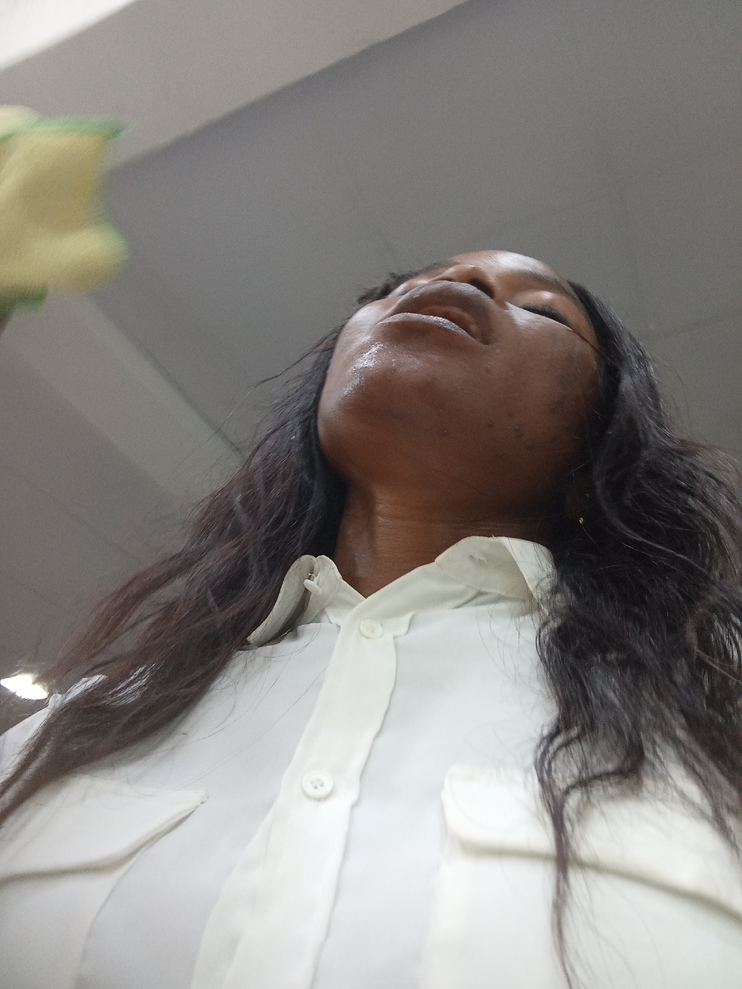 The image depicts a close-up, low-angle shot of a dark-skinned woman from the chest up. She is wearing a white collared button-down shirt. Her head is tilted back, exposing her neck and chin. Her eyes are closed, and her mouth is slightly open, with a sheen of moisture on her lips and forehead, suggesting sweat or exertion. Her long, dark hair is draped over her shoulders. In the upper left corner, a blurred object with yellow and green hues is visible, potentially a flower or a plant. The background appears to be an indoor environment, possibly a room with a neutral-colored ceiling and walls. The overall lighting is soft, highlighting the woman's features. The location in Kason, Nigeria, is not discernible from the visual information provided in the image.