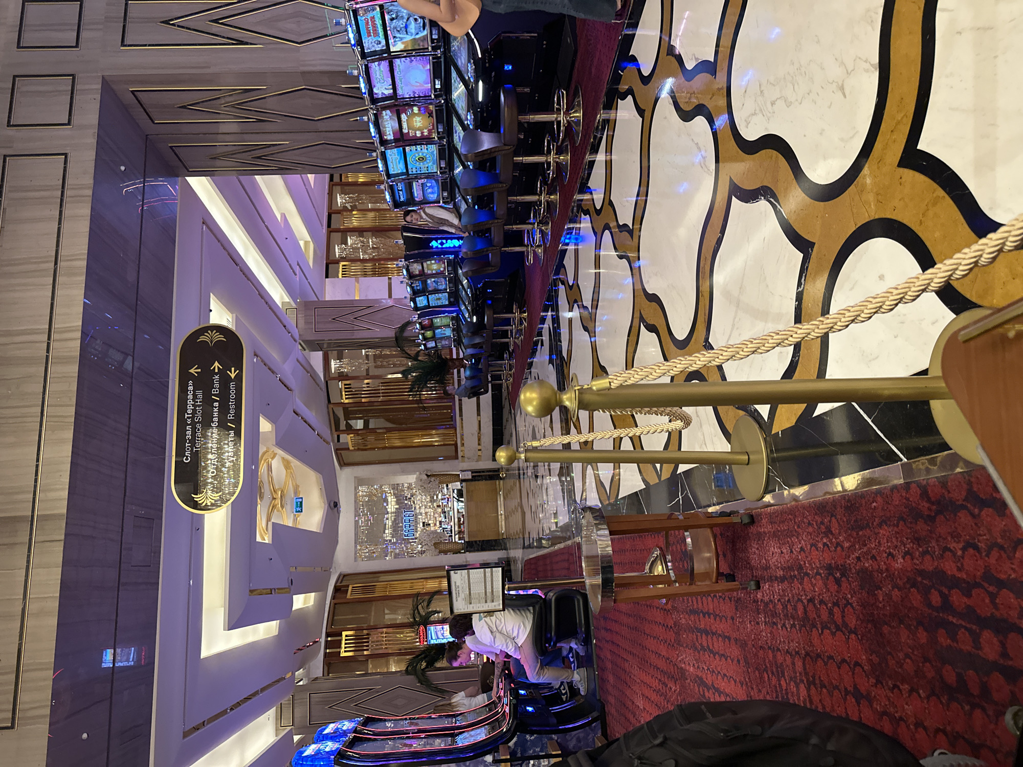 The image captures the opulent interior of what appears to be a casino or a high-end entertainment complex in Estosadok, Russia, as indicated by the visible text. The setting is characterized by luxurious decor, including polished marble floors with intricate gold and black patterns, rich red patterned carpeting, and walls adorned with gold trim and geometric designs.
In the foreground and middle ground, a row of modern slot machines with brightly lit screens displays various game interfaces. Several people are present; one individual wearing a white shirt and light pants is seated at a slot machine, engrossed in play, while another person is partially visible near the top of the frame. Golden stanchions connected by braided ropes delineate a pathway or section of the floor, adding to the upscale ambiance.
Notable details include the sophisticated lighting, featuring recessed ceiling lights and reflections that create a vibrant atmosphere typical of an entertainment venue. A large, ornate gold fixture resembling a chandelier is visible further back in the hall. Potted palm plants are strategically placed, enhancing the decor.
A prominent black oval sign with gold text and directional arrows provides information in both Russian and English. The visible text reads:
- Слот-зал «Терраса» / Terrace Slot Hall
- Отделение банка / Bank
- Туалеты / Restroom
This signage guides visitors to different areas within the complex, such as the "Terrace Slot Hall," a bank branch, and restrooms. The overall scene suggests a lively yet elegant environment where visitors engage in gaming and leisure activities.