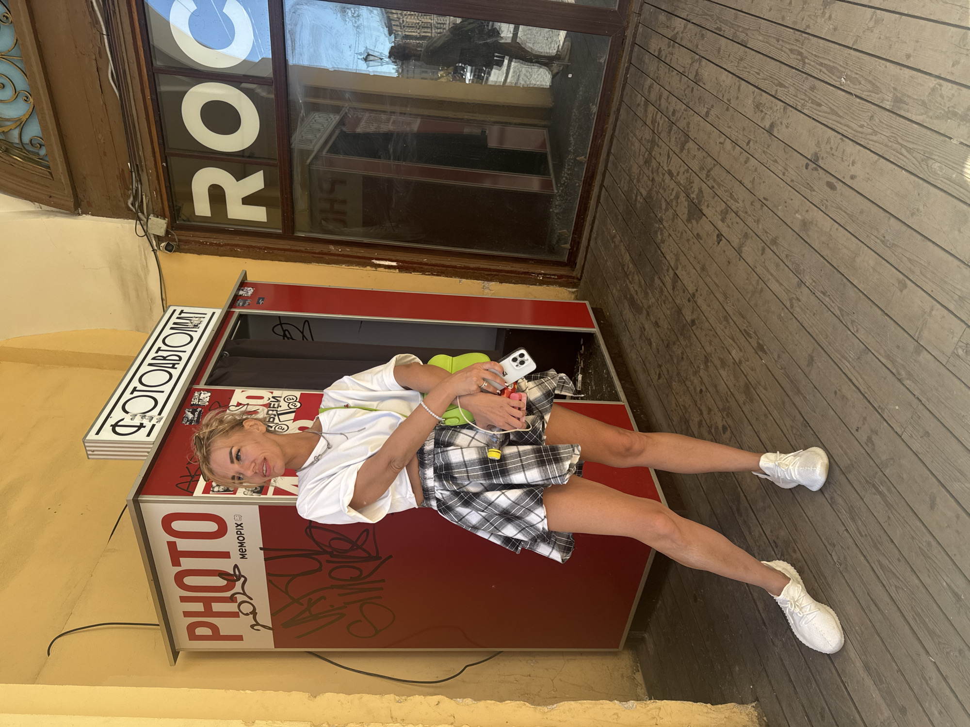 A young woman poses playfully in front of a red photo booth located on a wooden boardwalk. The woman is wearing a white t-shirt, a black and white plaid skirt, and white sneakers. She is holding a smartphone and a small green bag. Her blonde hair is pulled back, and she smiles at the camera.
The photo booth has "PHOTO" written in large red letters on its side, along with the brand name "MEMOPIX" and some graffiti. Above the booth, on the building, there is a sign with Cyrillic text that reads "ФОТОАВТОМАТ" (Photo Automaton). The background includes a window with large white letters spelling "ROC" visible through it, and reflections of buildings and street scenes. The setting appears to be outdoors, possibly in a public space in Saint Petersburg, Russia, given the context. The lighting suggests it is daytime, with clear, sunny weather. The woman's pose and expression convey a sense of fun and spontaneity.