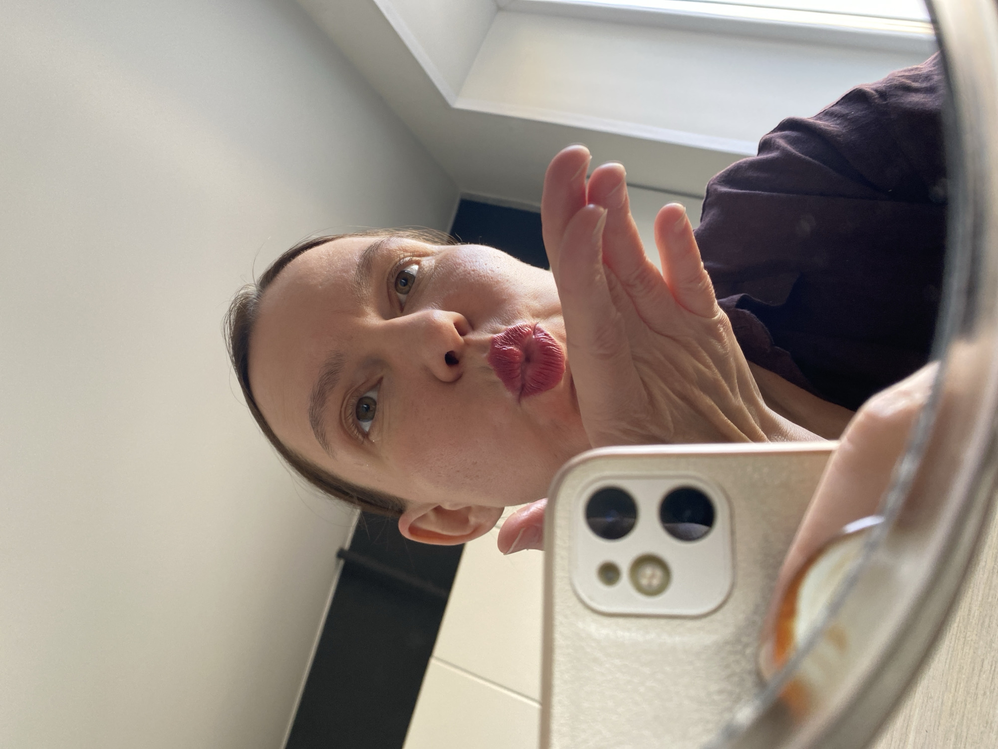 A woman is taking a selfie in a bathroom mirror. She is pursing her lips for the photo. The background includes a partially visible window and a wall with black and white tiles. She is holding a smartphone. The location is implied to be the city of Chaikovsky, Russia.