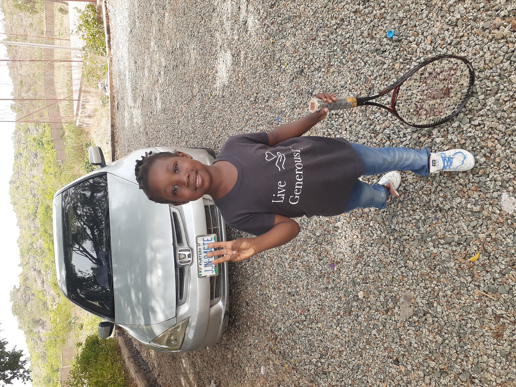 The image shows a young person standing next to a silver Honda car in an outdoor environment. The background suggests a rocky landscape, possibly in or near Jos, Nigeria. The person is holding a tennis racket and making a hand gesture. The car has a Nigerian license plate that says, "PLATEAU JUN-346TE". The ground is covered in small gravel. There's a blue plastic bottle cap near the tennis racket.