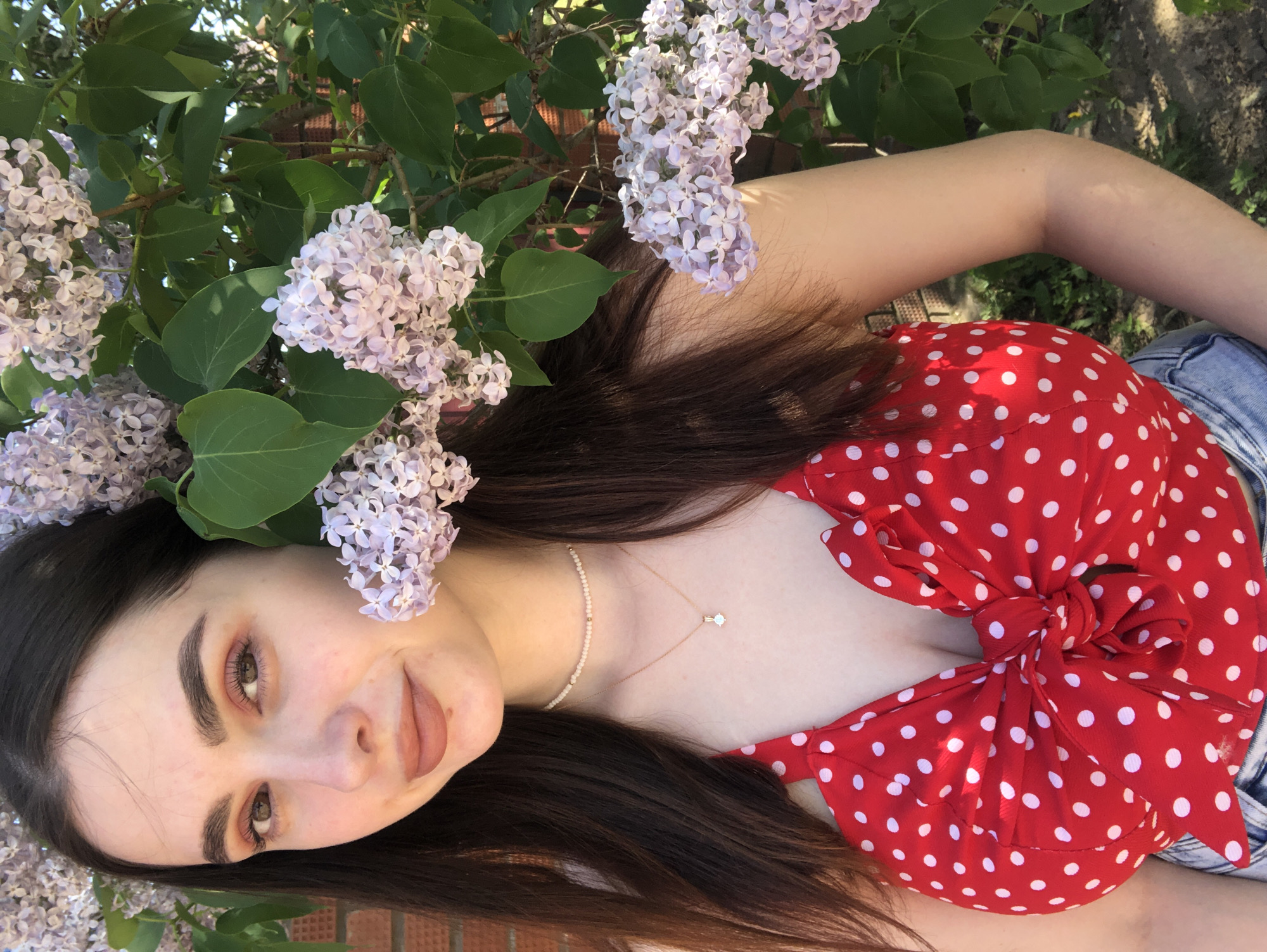 A young woman poses outdoors with lilac flowers. She has long, dark hair and wears a red top with white polka dots. She is wearing jewelry. Lilacs are visible behind and around her. The location cannot be determined.