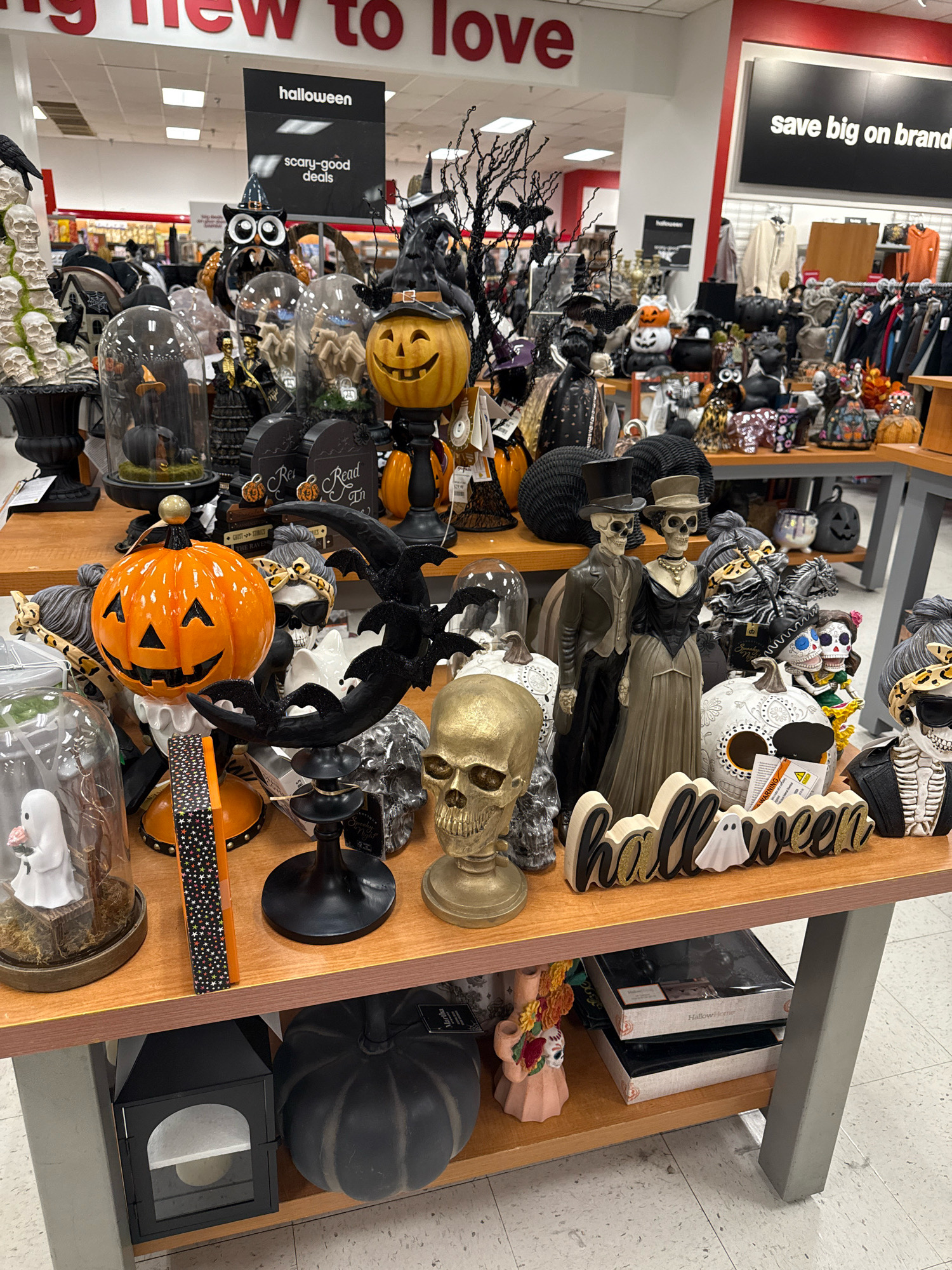 The image shows a retail store display of Halloween decorations in Commack, United States of America. The display features a variety of Halloween-themed items, including pumpkins, skulls, skeletons, ghosts, and decorative signs. The items are arranged on wooden shelves, with some items placed in glass domes. In the background, racks of clothing are visible, suggesting this is a department store or a retail chain store. Signs advertising "halloween" and "scary-good deals" are also displayed. The lighting is bright and even, typical of an indoor retail environment.