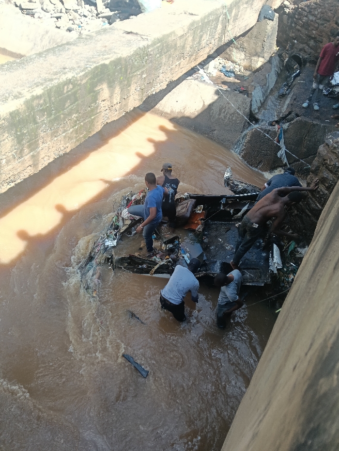 The image depicts a daylight scene in Nairobi, Kenya, centered on a recovery or salvage operation within a turbid, brown waterway. A severely damaged vehicle chassis, consistent with a large bus or truck frame, is partially submerged and entangled with debris, resting against a concrete bank on the right side of the frame. Seven adult males are directly observable in the scene, with shadows indicating additional individuals present.
Four men are waist-deep in the water around the wreckage: one man in a blue shirt and dark pants is seated on top of the vehicle frame; a second man, wearing a dark shirt and cap, stands behind him on the wreckage. To their right, two other men, wearing light-colored shirts and dark pants, are bent over, actively engaged with the submerged vehicle structure. A fifth man, shirtless, is positioned between the vehicle and the right concrete bank, appearing to push or pull. Two additional men are visible standing on the debris-covered bank to the right. Shadows consistent with human forms are cast upon the water and the left concrete retaining wall. The waterway flows from the upper left to the lower right of the frame, carrying visible refuse and organic debris. The banks on both sides consist of concrete structures and accumulated waste.