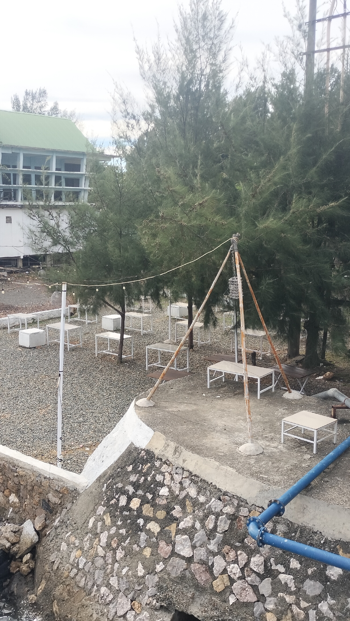 The image depicts an outdoor seating area with a cafe-like atmosphere, situated near a body of water. In the foreground, a stone-lined embankment slopes down towards what appears to be water, with a large blue pipe system running along the concrete edge. Behind this, a grey gravel-covered area is laid out with numerous white tables and some cube-shaped seats, arranged between several tall, slender trees. One prominent feature is a rustic, A-frame metal structure with a hanging lantern, adding a decorative element to the outdoor space.
In the background, a multi-story building with a green roof and a light-colored facade is visible. This building has a series of open balconies or walkways. The setting appears to be a coastal or waterfront location. The overall impression is that of a restaurant or cafe designed to take advantage of an outdoor, natural environment. The lighting suggests it is daytime, with the sky appearing overcast. There is no visible text in the image. The scene is devoid of people, giving it a sense of quietude. The abundance of trees and the proximity to water suggest a potentially temperate or tropical climate. The presence of cafe furniture implies a leisure or dining establishment.