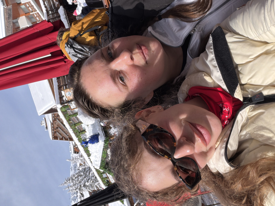 Two females are photographed outdoors in a snowy alpine environment, specifically in the city of Les Allues, France. The foreground features the upper torsos and faces of two individuals.
The person on the left has light brown, curly hair and wears dark tortoise-shell cat-eye sunglasses with gold accents on the temples. She is dressed in a light-colored puffer jacket, possibly cream or off-white, with a red bandana visible around her neck. Her lips are slightly parted in a subtle smile.
The person on the right has dark brown hair, pulled back, and discernible blue eyes. She wears a light grey or white ski jacket over a dark layer and looks directly towards the camera with a neutral expression.
The background consists of multiple snow-covered wooden chalet-style buildings, characteristic of a ski resort. These buildings have snow-laden roofs and balconies decorated with green garlands and string lights. A blue decorative object rests on a snow-covered ledge of one building. Snow-covered evergreen trees are visible on the mountain slopes behind the structures. Portions of large red and black outdoor umbrellas are also present. The sky is clear blue, indicating a sunny day. Distant figures in winter clothing are visible further in the background, suggesting an active outdoor area.