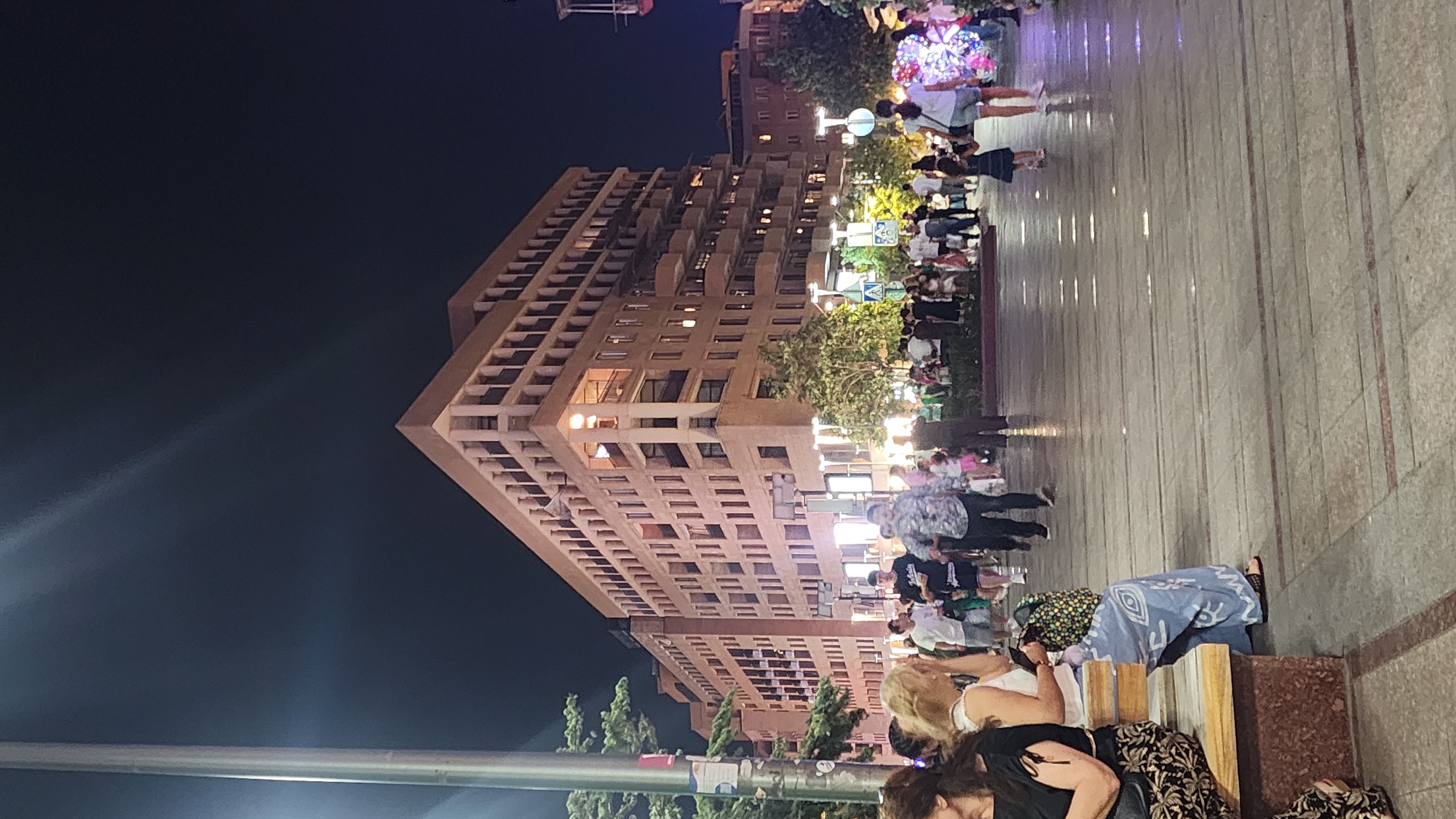 This is a nighttime, vertical-format photograph taken on a city street in Yerevan, Armenia. The scene depicts a busy pedestrian area with a large, multi-story building dominating the background. The building is light brown and has numerous balconies and windows, with some lights on inside. The street in the foreground is paved with large square tiles and appears wet, possibly from recent rain, reflecting the lights from the building and surrounding area.
There are many people walking and standing on the street. In the foreground, two women are sitting on a bench. One is wearing a black top and a patterned skirt, and the other is wearing a white top and a large blue patterned garment. Several other individuals are walking in the mid-ground, including a man in a patterned shirt and shorts, and another man in dark clothing. Further back, a crowd of people are gathered, some appearing to be looking at something. There are also trees and streetlights visible, contributing to the illuminated atmosphere of the night scene. The overall impression is one of a lively urban environment in Yerevan.