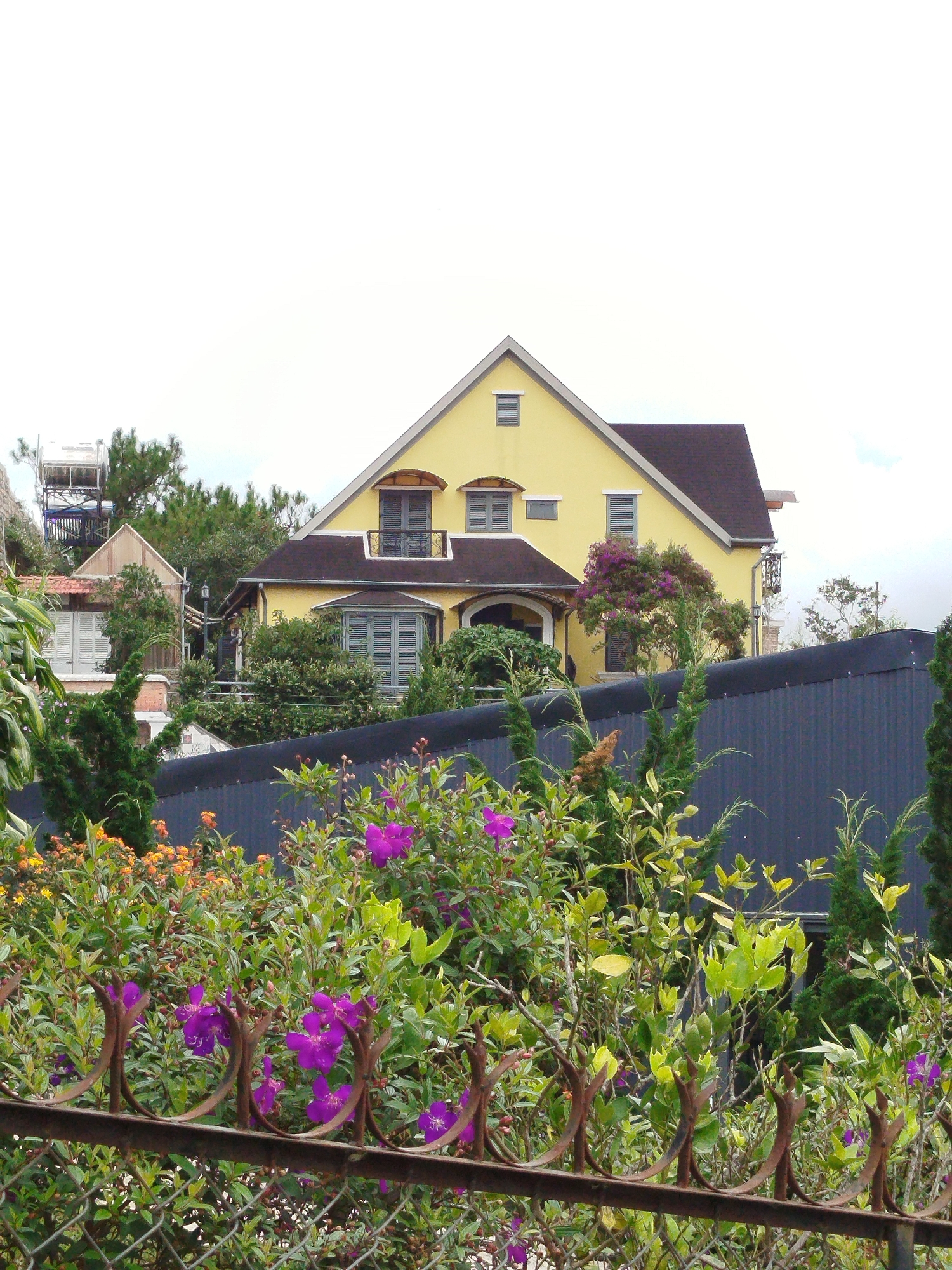 In the city of Da Lat, Vietnam, a bright yellow house with a dark brown roof is situated on a slope. The house features multiple windows, some with shutters, and a prominent pitched roof. In the foreground, a dense arrangement of green foliage and vibrant purple and orange flowers is visible behind a rusty, decorative metal fence and a chain-link fence. The background includes a dark, corrugated metal wall and various trees, with a water tower on a metal frame visible to the left of the house. The sky is overcast.