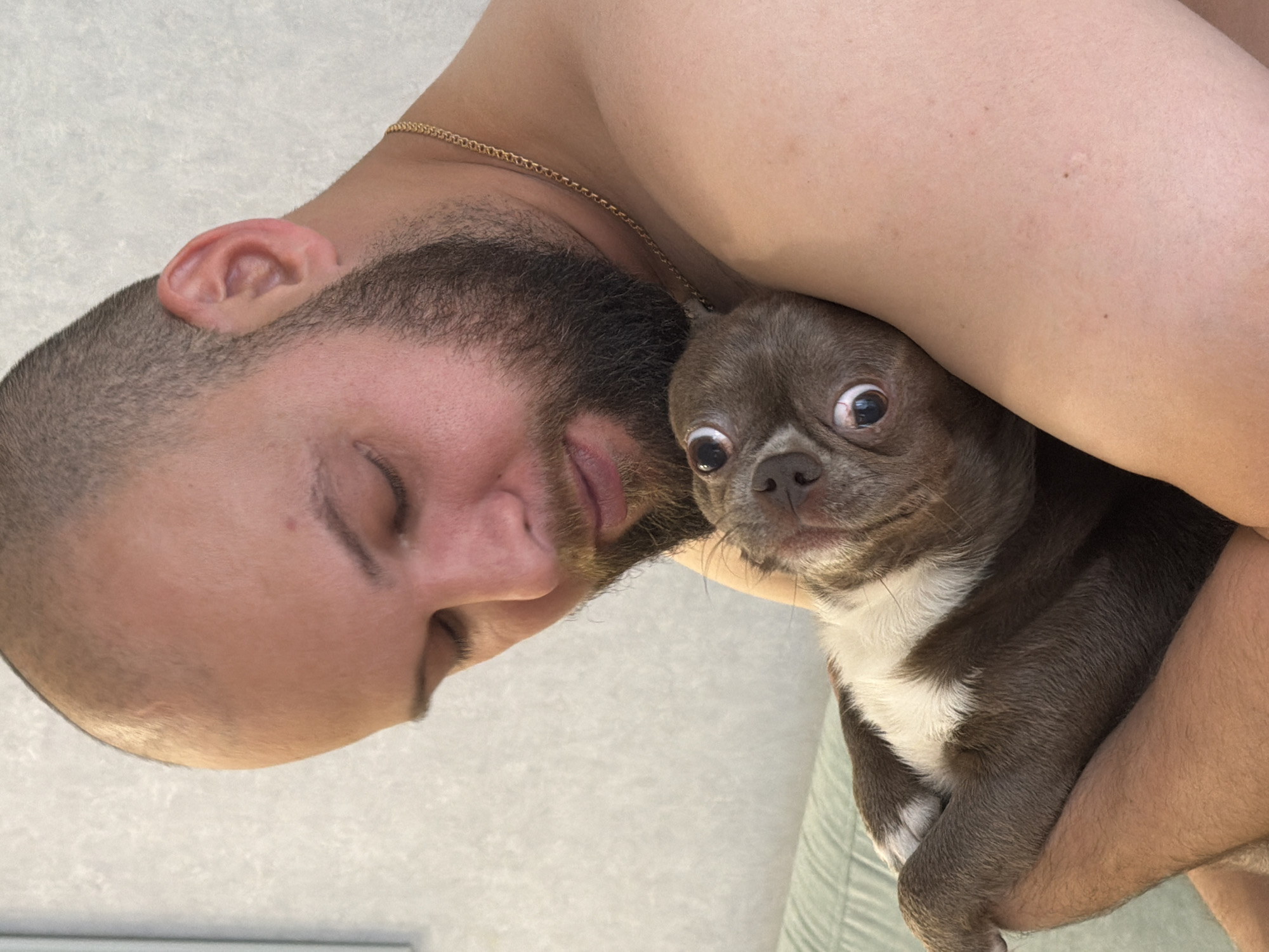 This media file features a close-up shot of a person and a dog. The person, with short hair and a beard, is lying down, with their head tilted to the side, and eyes closed. They are wearing a gold chain around their neck. The dog, a small breed with brown and white fur, is being held by the person, with its head peeking out and wide eyes looking directly at the camera. The background appears to be an indoor setting, possibly a bedroom or living room, with textured walls. The lighting suggests it might be daytime, but there are no clear indicators of the time of day or weather. The overall mood seems intimate and relaxed, with the person holding the dog in a close embrace. No text is visible in the image. The context of Votkinsk, Russia, does not provide any further visual cues within the image itself.