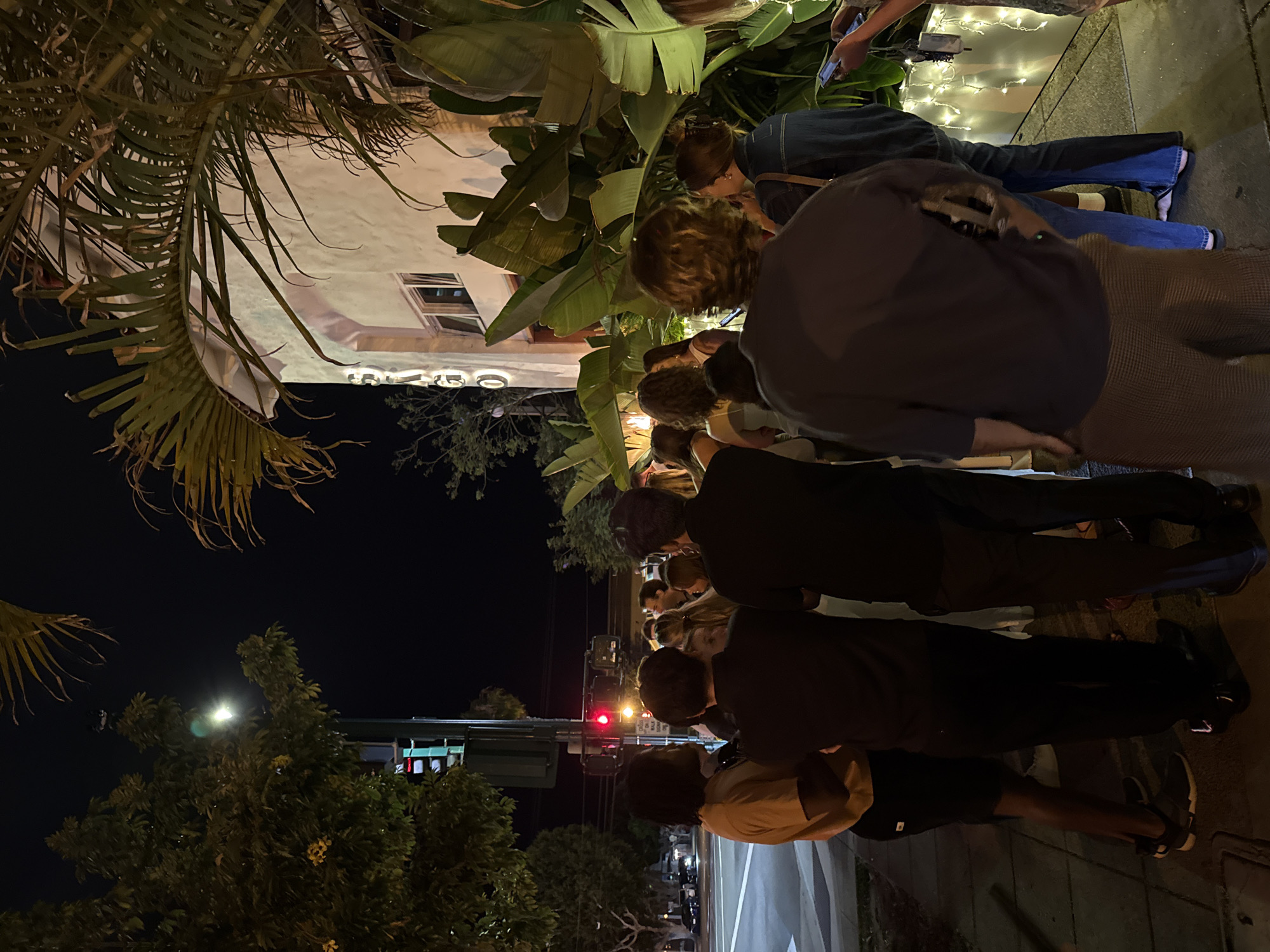 A group of people stand on a sidewalk at night in front of a building. The building appears to be a restaurant or bar, with large tropical plants in front. The street and sidewalk are wet, suggesting it may have recently rained. A traffic light is visible in the background, with a red light illuminated. The people are casually dressed and appear to be waiting in line or socializing. One person in the foreground is looking at their phone. The overall atmosphere is relaxed and social. The location is on South Coast Highway 101 in Encinitas, California, United States of America. The name "SANTA" is visible on the building's facade.