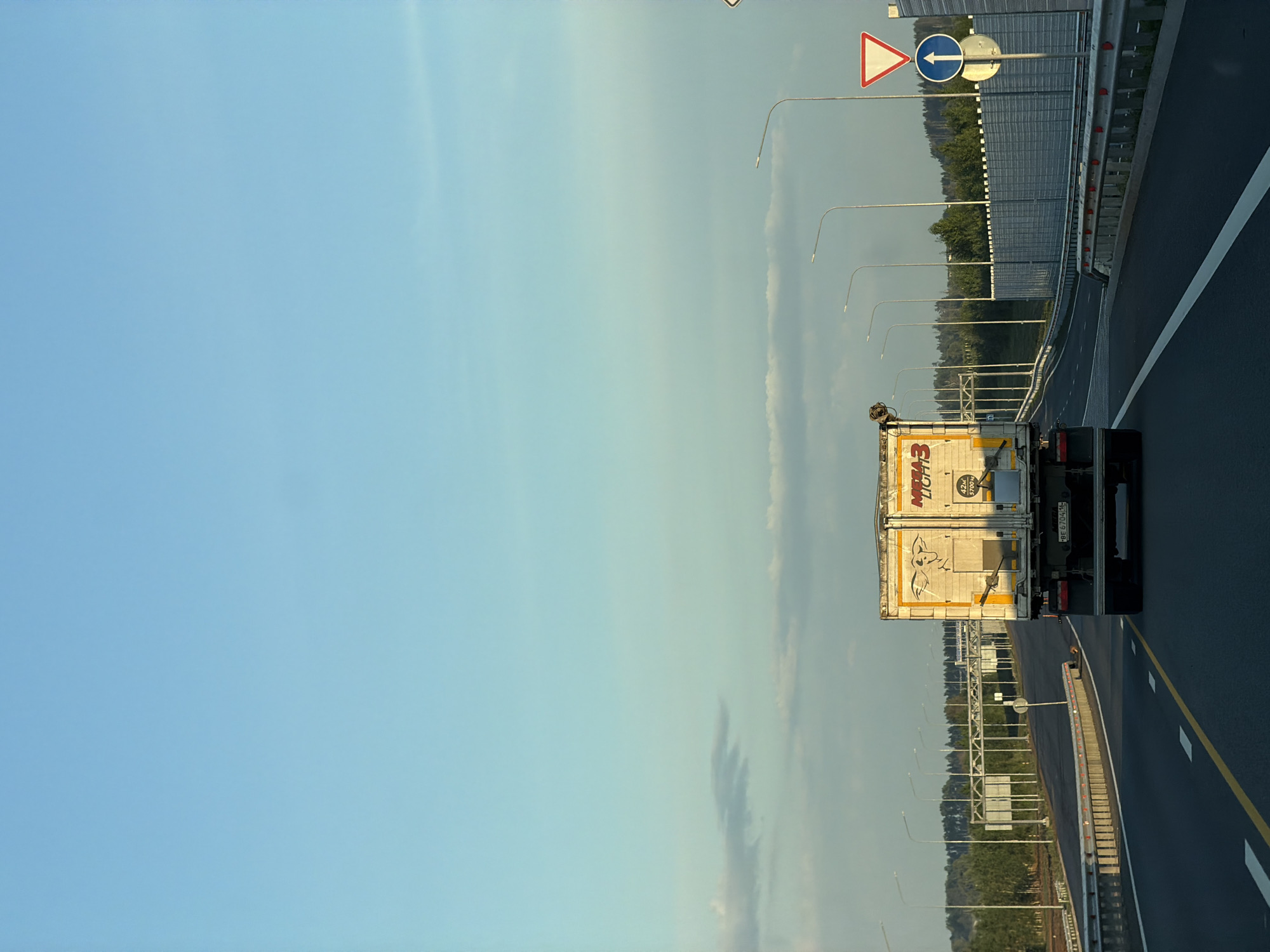 A truck is driving down a highway in Kaipy, Russia. The sky is blue with some clouds. The truck is white and has writing on the back. There are trees and other signs along the side of the road.