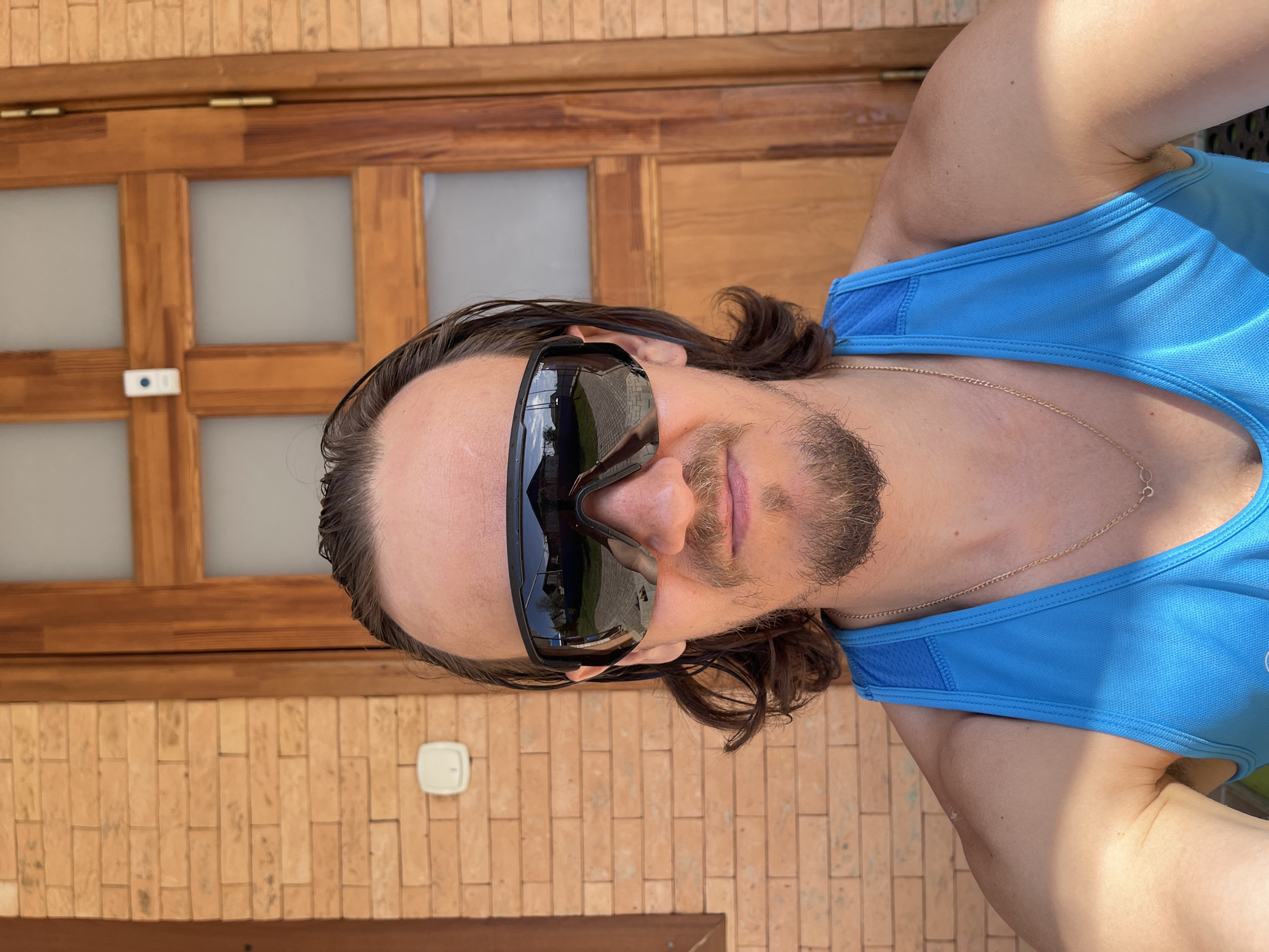 A man with long brown hair, wearing black sunglasses and a blue tank top, is facing the camera. He has a light beard and a gold chain around his neck. The background consists of a brick wall and a wooden door with glass panes. The location is Перевозное, Russia.