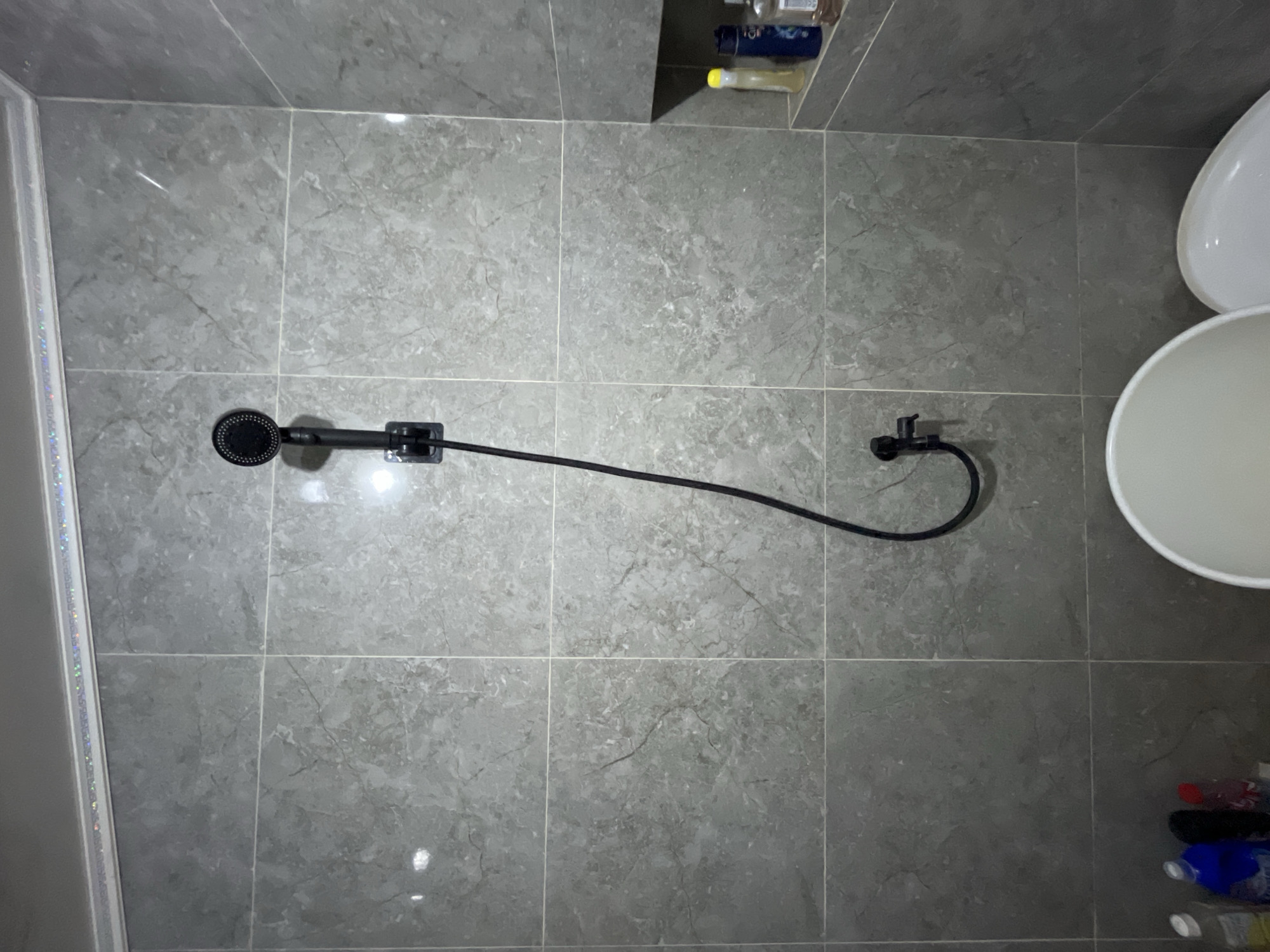 The image shows a close-up of a shower area in a bathroom. The walls are tiled with large, gray, marble-patterned tiles. A black showerhead is mounted on the wall, connected to a black flexible hose that curves downwards. A black faucet is attached to the hose near the bottom. In the background, on a small shelf, are several bottles of toiletries, including a yellow bottle, a blue bottle with a white cap, and a clear bottle with a white cap. In the foreground, the rim of a white bucket and a portion of a white toilet seat are visible. The overall impression is of a functional but basic bathroom. The location of the image is stated to be in the city of East Jakarta, Indonesia.