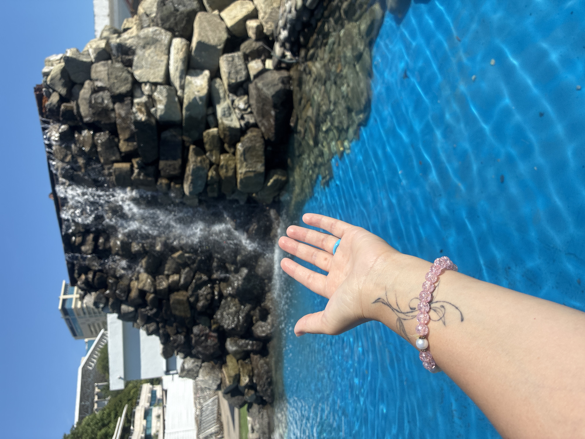 A person's left hand is held out over a bright blue pool of water, near a rock formation with a small waterfall. The hand has a tattoo on the wrist of a swirling vine design and is adorned with a pink beaded bracelet with a pearl. A bright blue ring is visible on the index finger. The background shows buildings and trees under a clear blue sky. This scene is located in Sochi, Russia, likely within a resort or park area.