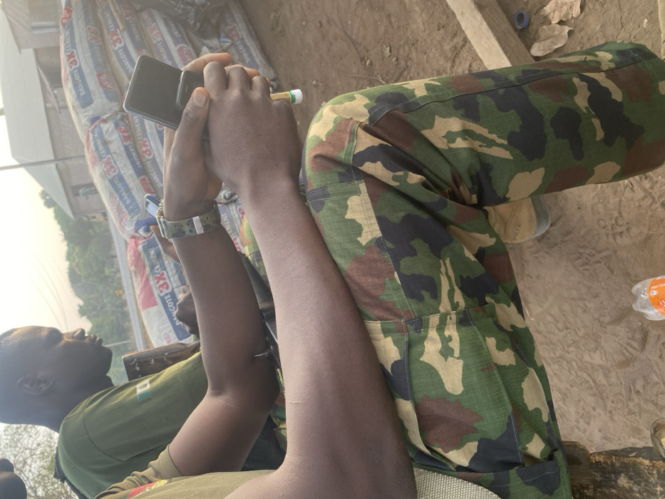A person wearing camouflage pants and a green shirt is holding a phone in their hands. The person also wears a camo watch on their wrist. They appear to be sitting down. Behind the person are stacked bags labeled "DANCOTE 3X CEMENT". To the side of the person are visible patches of ground and what appears to be a water bottle.