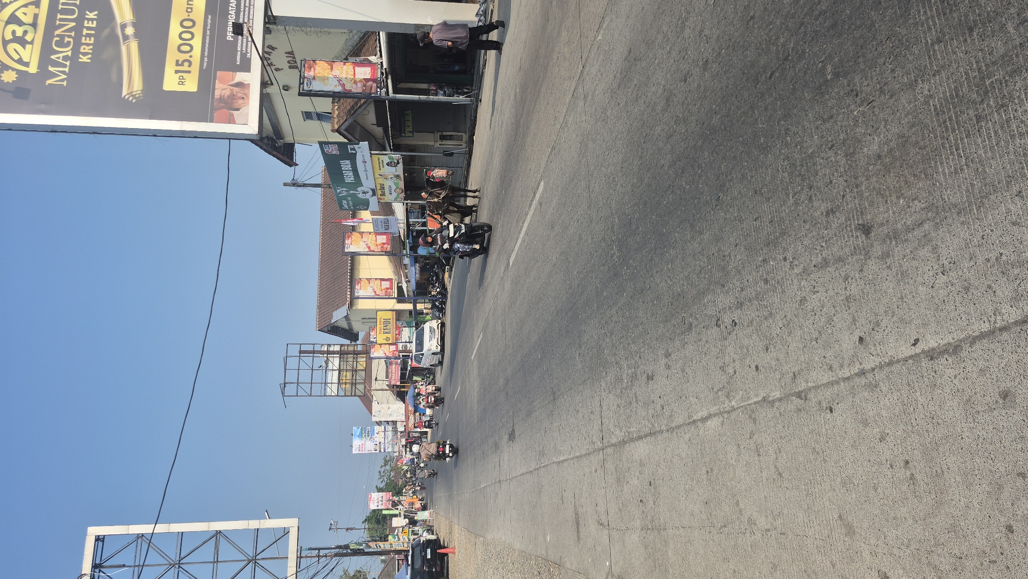 The image shows a street scene in Boja, Indonesia. The street is paved and has multiple lanes, with vehicles and pedestrians present. On the right side of the street, there are several storefronts and buildings, including "Pasar Boja" which appears to be a market. There are also billboards and signs advertising various products and services, including "Magnum Kretek". People are seen riding motorcycles and walking, and there is a horse-drawn cart with a person on it. The environment appears to be an urban or semi-urban area, with buildings lining the street and a clear blue sky overhead. The lighting suggests it is daytime.