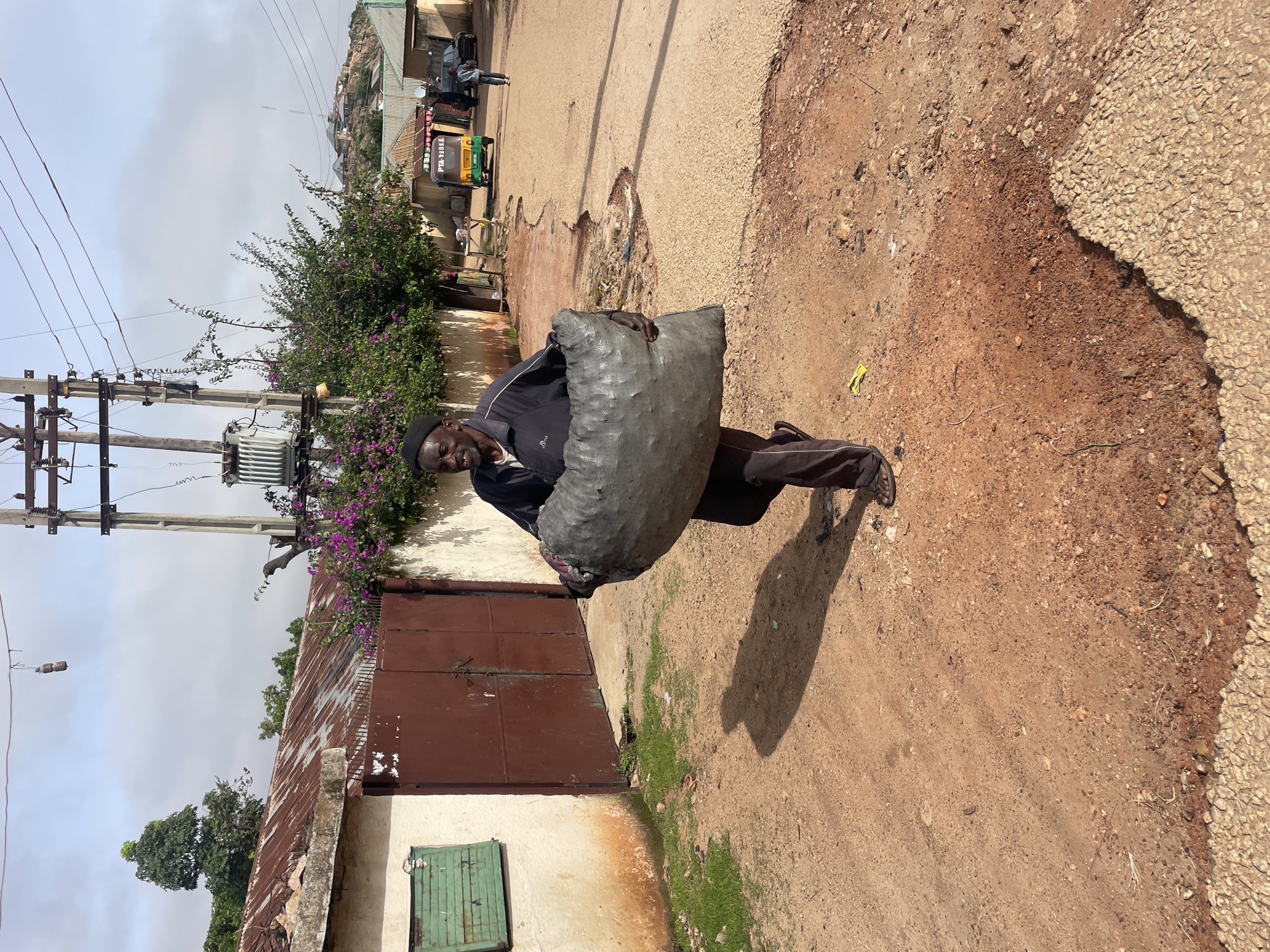 A man walks carrying a large sack of coal down a dirt road in the city of Jos, Nigeria. The man wears a black cap, a dark jacket, and dark pants with sandals. He is seen from the waist down. The sack of coal is gray and appears to be made of a rough material. The road is unpaved and reddish-brown, with some gravel and patches of green grass. In the background, there is a building with a rusty corrugated metal roof and a large red metal gate. Electrical poles and wires are visible overhead. Further in the distance, there are more buildings, a yellow and green auto rickshaw, and a black car. The sky is partly cloudy.