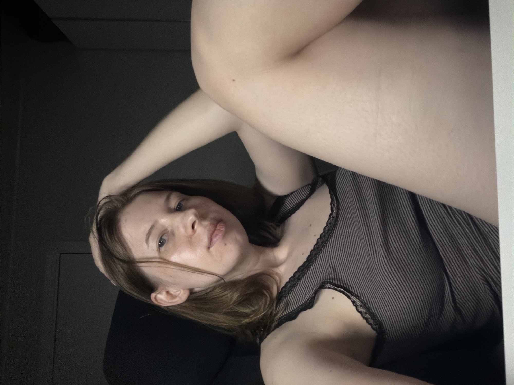 In a dimly lit room, a young woman with light brown hair is captured in a close-up, reclined pose. She is wearing a dark, pinstriped tank top with lace trim. Her right arm is resting behind her head, and her left leg is bent and crossed over her right. The background consists of dark walls and furniture. The lighting suggests an indoor setting, possibly a bedroom or living area, with the primary light source coming from the front, illuminating her face and upper body. The composition is intimate, focusing on the woman's facial features and the curve of her body. There are no discernible landmarks or elements that would indicate a specific location within Dongducheon-si, South Korea.