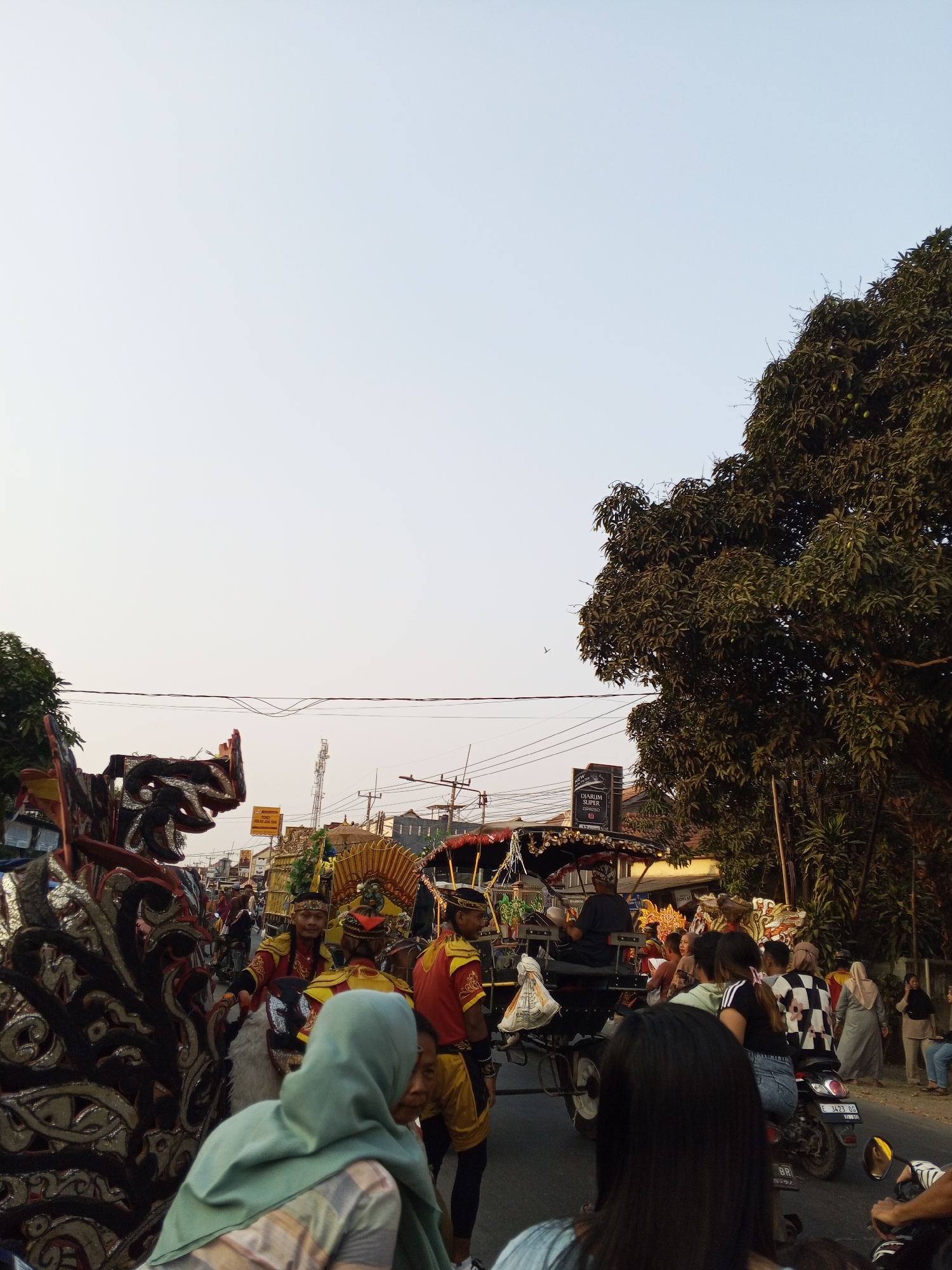 A street scene in Gebang, Indonesia, features a parade or festival. A large, ornate dragon-like float is prominent on the left, adorned with intricate black, gold, and red designs. Several individuals in traditional, colorful costumes, likely performers, are visible around the float and in the background. Some are wearing red and gold attire with headbands, while others are dressed in yellow and red.
A horse-drawn carriage, decorated with gold accents and a canopy, is also part of the procession. People are gathered on the sides of the street, observing the event. A motorcycle with two passengers, one wearing a checkered shirt, is in the middle ground. A large tree with lush green foliage dominates the right side of the frame, casting some shadows. Power lines and poles are visible in the background, indicating an urban or semi-urban setting. The sky is clear and pale blue, suggesting it is daytime, likely late afternoon or early morning due to the warm light.