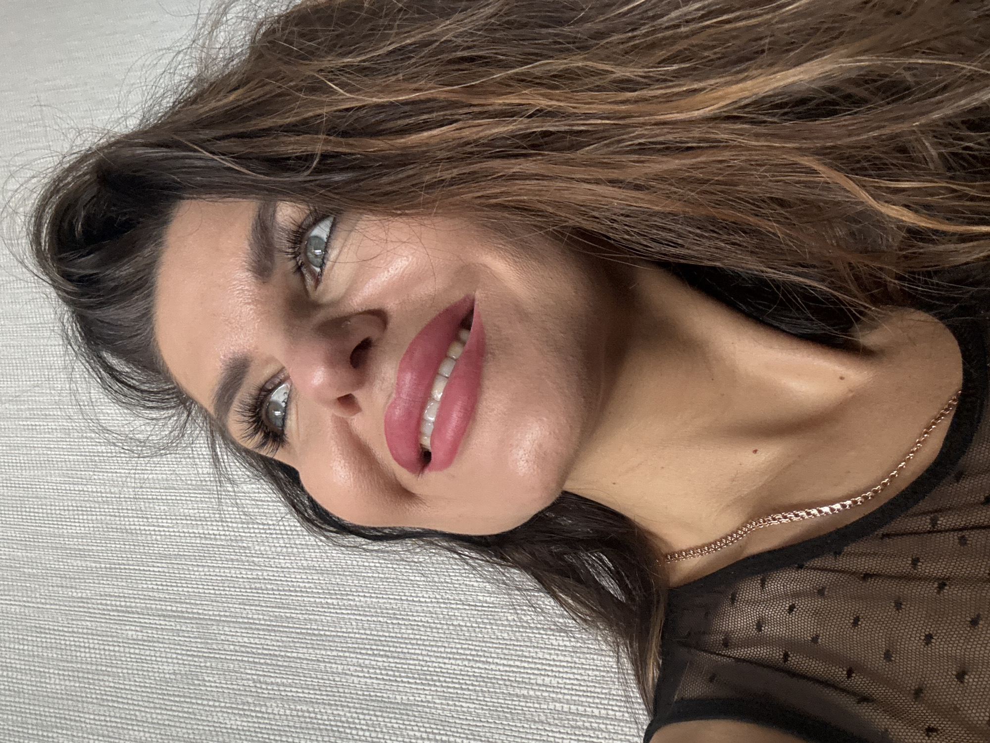 A close-up, rotated portrait shows a woman with long, wavy brown hair highlighted with blonde streaks. Her skin is fair, and her eyes are light-colored, possibly gray or blue. She has full lips coated in pink lipstick and visible white teeth. A delicate gold chain necklace adorns her neck. She is wearing a semi-sheer black top with a subtle pattern. The background is a textured gray surface. The image provides no information to determine a location or a connection to the city of Dubovaya, Russia.