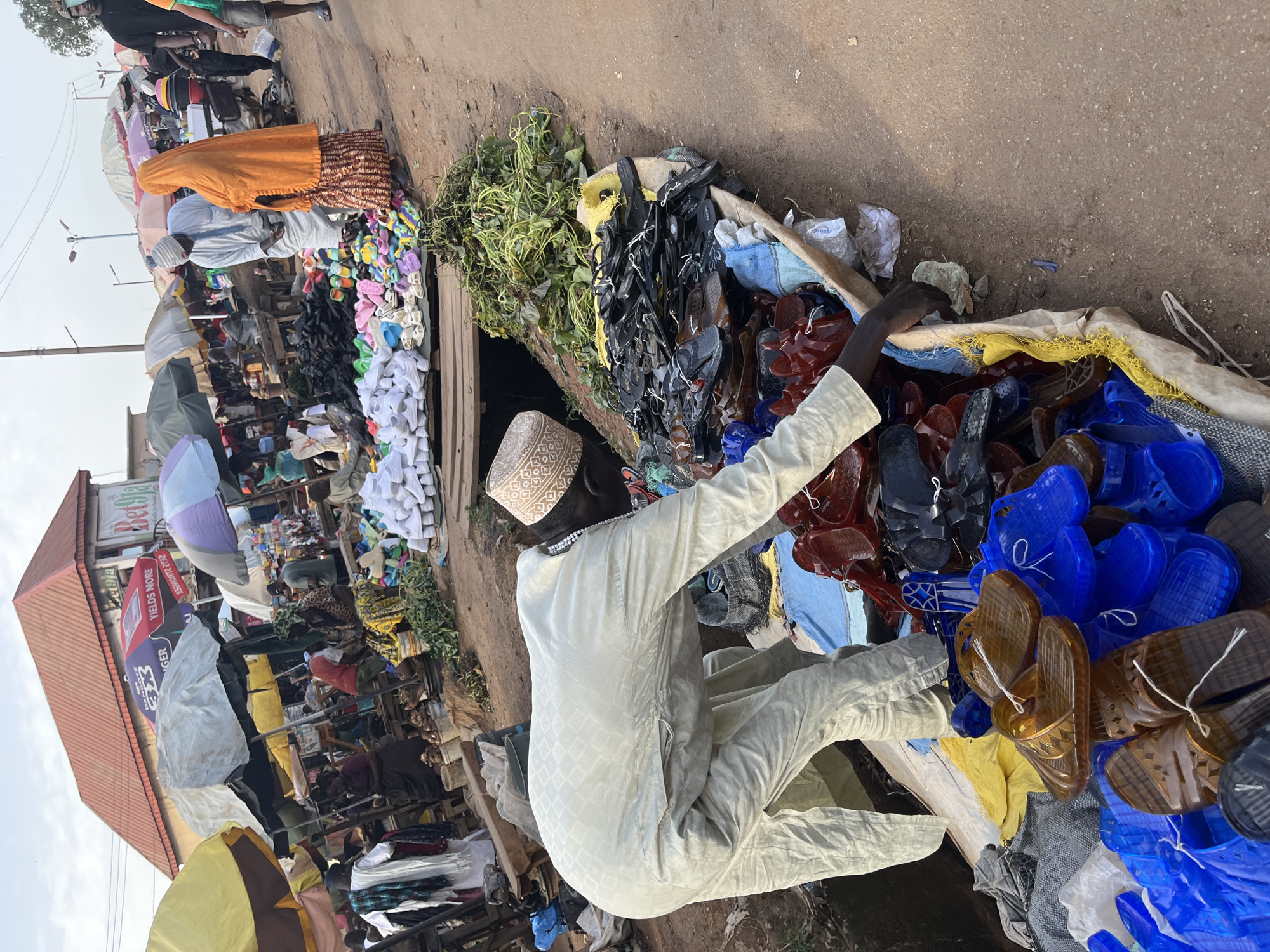 This is a street market scene in Rafin Par, Nigeria. A man wearing a light-colored traditional outfit and a patterned cap is bending over a stall, appearing to sort or display a large quantity of footwear. The stall is overflowing with various types of sandals and flip-flops, predominantly in black, brown, and blue colors. Some are made of rubber and plastic, while others are a lighter material, possibly white canvas shoes are also visible in the background. The market is bustling with activity, indicated by the presence of other people in the background, some of whom are also dressed in traditional Nigerian attire. Several stalls with umbrellas offering shade are set up, selling various goods. The ground is a mix of dirt and paved pathways, suggesting an outdoor market environment. The architecture in the background includes a red-roofed building with signage that appears to be in a local language. The overall impression is of a vibrant and busy marketplace.