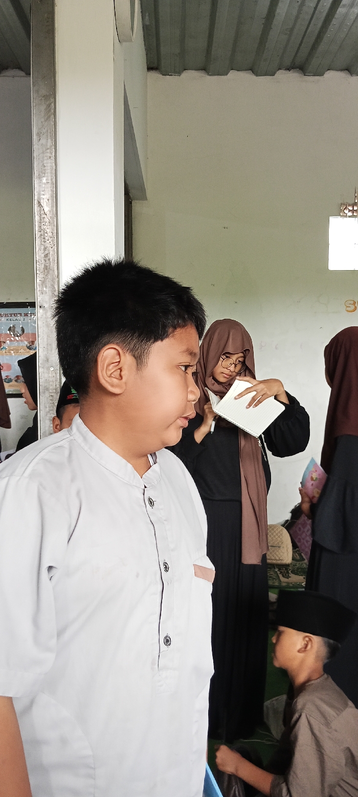 The image shows a young boy in a white collared shirt standing in the foreground. He is looking off to the right. To his right, a woman with long brown hair is wearing a brown hijab and black clothing, holding a notebook. She appears to be looking down at the notebook, her expression neutral. She is standing to the right of the boy. In the background, there is a young boy kneeling on the floor. He is wearing a brown shirt and black hat and is looking towards the woman. The environment appears to be an indoor space with white walls and a metal roof. It is not possible to say what the boy is doing. The location, based on the coordinates provided, is in the Cangkuang area of West Java, Indonesia.