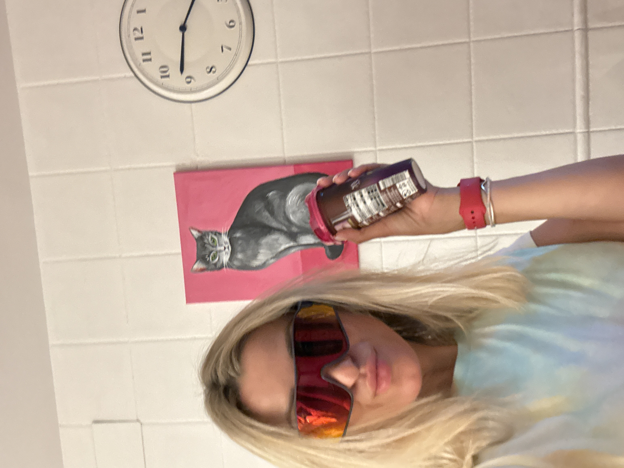 A blonde woman wearing oversized, reflective sunglasses and a tie-dye t-shirt is holding a purple bottle with a red lid. She is standing in front of a tiled wall, to the left of a painting of a grey cat on a pink background. A round clock is visible on the wall to the right of the cat painting. The woman is also wearing a pink smartwatch and a silver bracelet. The scene appears to be indoors, likely in a bathroom or kitchen based on the tilework. There is no indication of the specific location within the city of València, Spain.