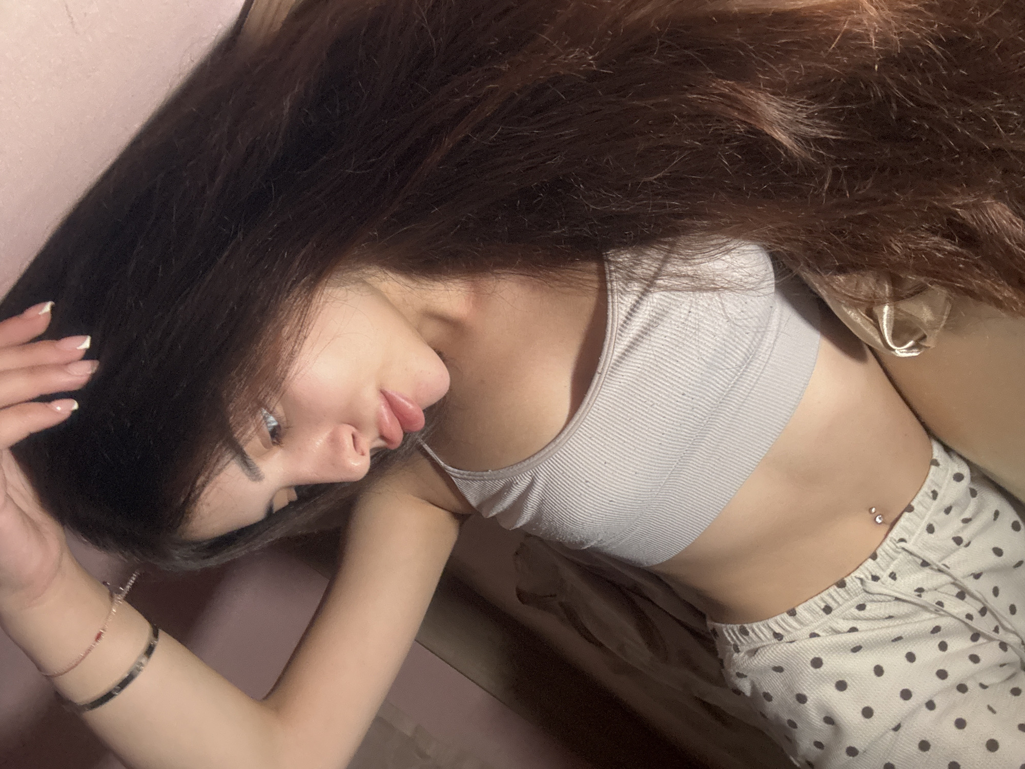 A young woman is depicted lying down indoors, likely on a bed or sofa, in Plovdiv, Bulgaria. She has long, flowing dark brown hair that spreads upwards in the frame. She is wearing a light grey ribbed sports bra or crop top and light-colored pajama pants with dark polka dots.
Her left arm is bent, and her hand, adorned with a French manicure, rests near her head. Two bracelets are visible on her left wrist: a delicate chain with small red accents and a wider, solid dark band. Her face is shown in profile, revealing her left eye, nose, and lips, which curve into a gentle, relaxed smile. A silver barbell belly button piercing with two gems is visible on her midsection.
The background primarily features a plain light pink or peach-colored wall on the left and a darker, possibly beige, fabric surface on the right. The image captures a casual and relaxed moment. There is no visible text in the image.