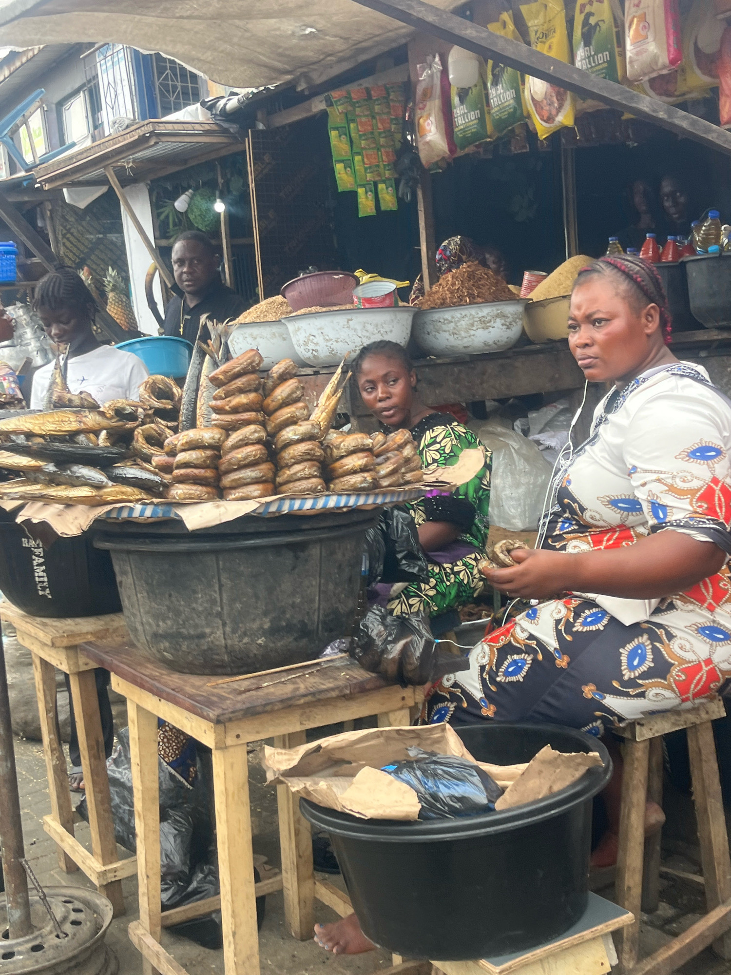 In Lekki Phase I, Nigeria, a bustling market scene unfolds under a makeshift canopy. Three women are visible, engaged in commerce. On the right, a woman wearing a white, red, and blue patterned dress with blue eye-like motifs, and earphones in her ears, is seated and holding a piece of fish. To her left, another woman in a green and black patterned dress is also seated, looking towards the left of the frame. A third woman, wearing a white t-shirt, is standing and looking down.
The foreground features several large, dark tubs and wooden crates, supporting a variety of goods. A prominent display of smoked fish is stacked in rows on a blue and white striped cloth covering a large tub. Other tubs contain dried goods, appearing to be grains or legumes, and a large black tub in the lower right corner holds discarded wrappers and a dark plastic bag. The background reveals more market stalls with shelves stocked with various items, including bags of what appears to be rice and other packaged goods. The overall environment suggests a vibrant, open-air market with a focus on food sales, particularly fish.