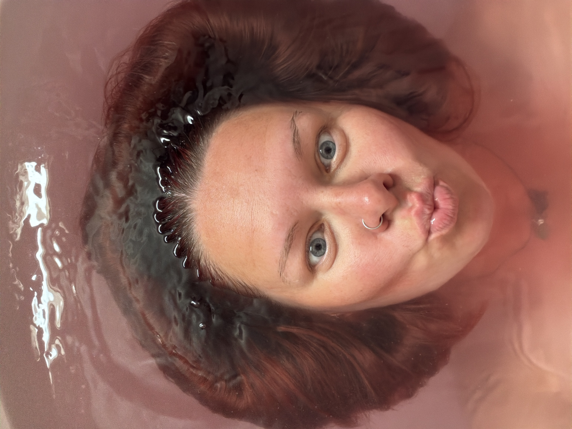 A woman is submerged in a pinkish-colored bathwater, her face and the top of her head visible above the surface. She has fair skin, light blue eyes, and pursed lips. A silver ring is visible in her left nostril. Her brown hair is partially submerged in the water, appearing darker where wet. The water around her head has ripples and reflections, suggesting movement. The location is indeterminate and cannot be identified from the image. The city of Chaikovsky, Russia is not relevant or identifiable in this image.