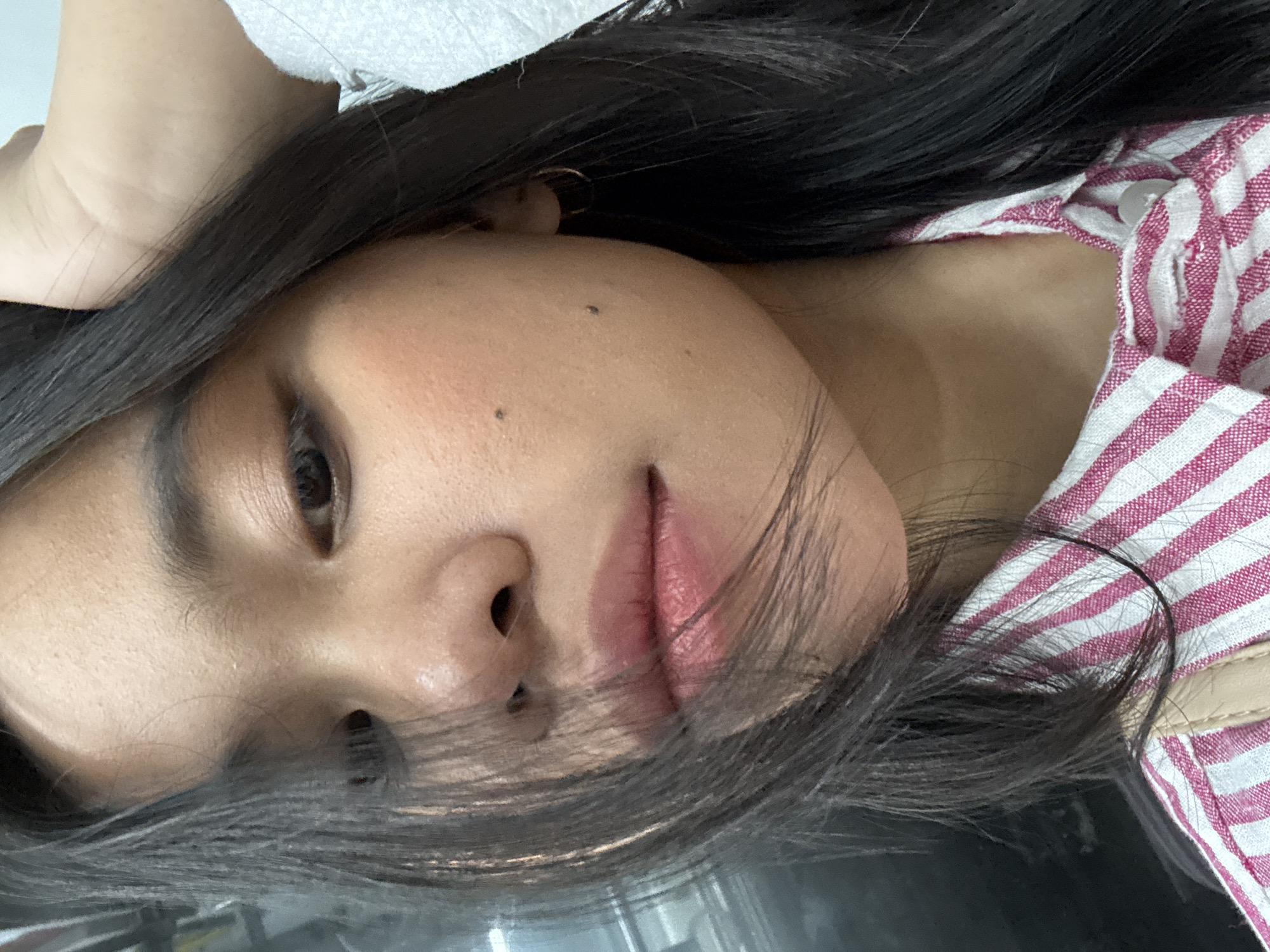 The image is a close-up, vertically oriented selfie of a young woman. She has long, dark hair that falls across her face and shoulders, obscuring parts of the frame. Her head is tilted to the side, and her eyes are looking towards the camera. She is wearing a pink and white striped shirt, and a light-colored fabric is visible in the upper left corner of the image. One of her hands appears to be resting on her head. The woman has on makeup, including what appears to be lip gloss and blush. There are a couple of small moles visible on her face. The lighting suggests it is daytime, possibly indoors, given the quality of the light. There are no visible text elements in the image. The overall impression is a casual, personal snapshot.