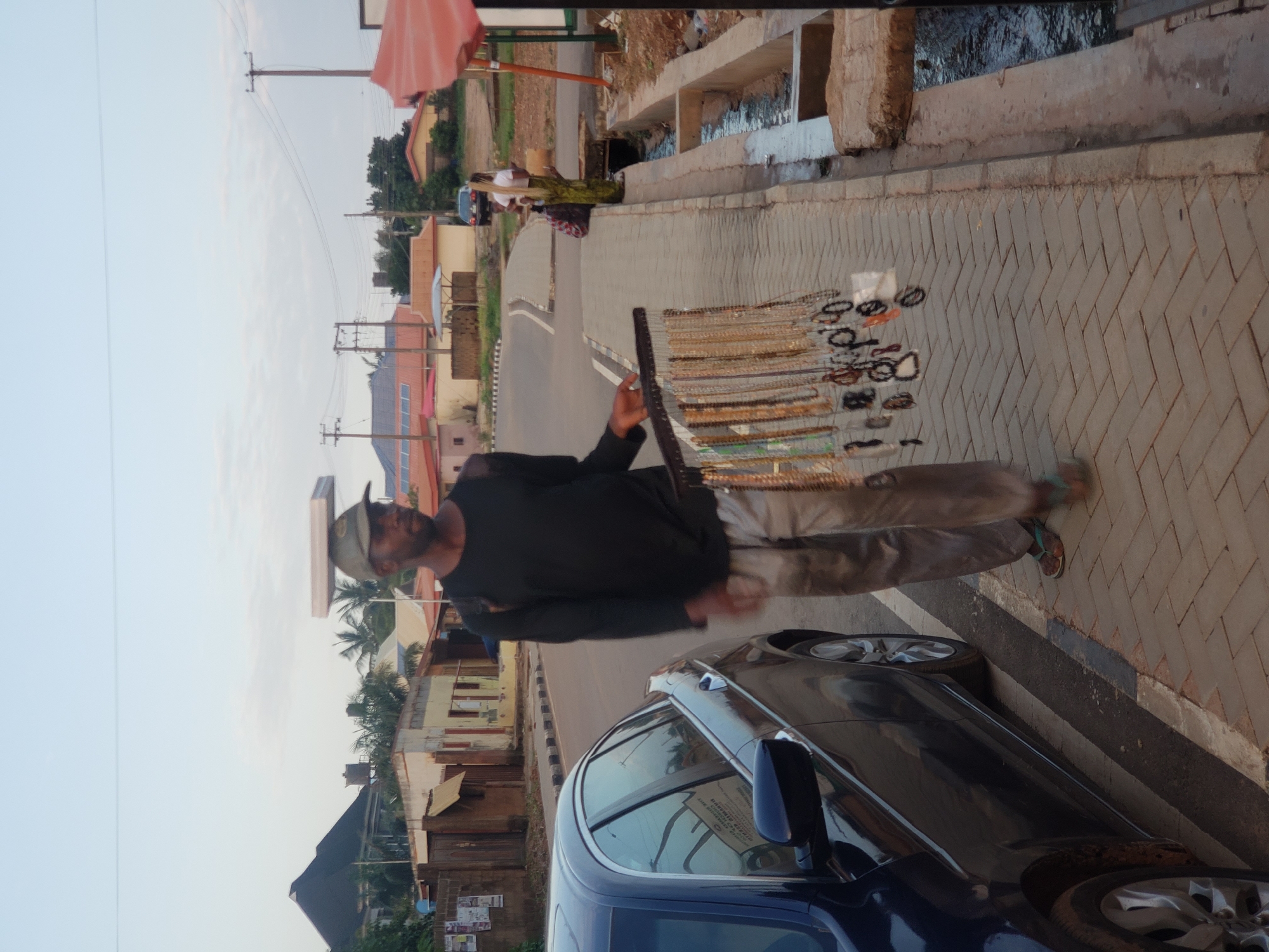 A man wearing a dark long-sleeved shirt, light-colored pants, and flip-flops walks along a paved sidewalk in Narayi, Nigeria. He carries a display rack of necklaces and jewelry on a stick balanced horizontally. A flat, light-colored object is balanced on top of his baseball cap. A dark blue sedan is parked on the street next to the sidewalk. Buildings with red and orange roofs line the street in the background, along with utility poles and wires. A narrow concrete channel with water runs alongside the sidewalk. The sky is overcast.