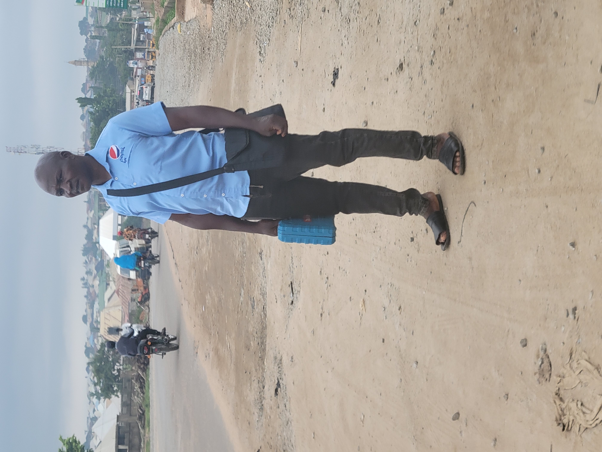 An image captures a full-body shot of a man standing on an unpaved road in what appears to be a semi-urban or rural setting in Suleja, Nigeria. The man, with a bald head and a neutral expression, looks directly at the viewer. He wears a light blue polo shirt featuring the "Pepsi" logo on the left chest, dark trousers, and dark sandals. A black shoulder bag crosses his torso, and he holds a small, rectangular blue object in his left hand, down by his side.
The road he stands on is sandy/dusty with scattered small stones, and it appears to be a shoulder or side lane next to a slightly more defined but still unpaved main road. In the background, there is a busy scene with multiple motorcycles, some carrying passengers, and what looks like a few cars. Buildings, ranging from single-story structures to more substantial ones, are visible along the roadside and stretching into the distance. A tall, light-colored structure, possibly a mosque or tower, is visible among the buildings. The sky is overcast or hazy, suggesting a bright but not sunny daytime. The overall impression is one of everyday life in a bustling local environment.