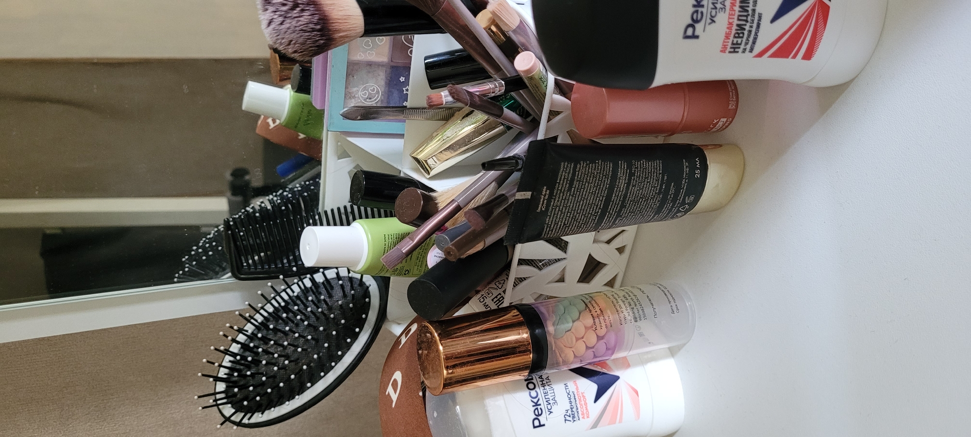 Here is a detailed description of the image:
**Scene:** A close-up shot of a collection of beauty and personal care products housed in a white storage container. The items are tightly packed and arranged at various angles.
**Objects:**
* Makeup brushes: Numerous makeup brushes with various sizes and types of bristles are visible.
* Cosmetics: There are tubes and bottles of cosmetic products, including foundation, primer, and possibly concealer.
* Hairbrush: A black hairbrush with white-tipped bristles.
* Deodorant: Two deodorant bottles are present. One is white with red and blue accents, the other is white and brown.
* Storage container: The items are organized within a white container with a geometric cutout design.
* Mirror: A mirror in the background shows a partial reflection of the surroundings.
**Environment:** The scene is indoors. The surface appears to be a white countertop or vanity.
**Text/Labels (OCR):**
* Labels in Russian language are present on the deodorant bottles: "Рексом" (Rekson), "УСИЛЕННА ЗАЩИТА" (Enhanced Protection), "АНТИБАКТЕРИА НЕВИДИМ" (Antibacterial Invisible), "НА ЧЕРНОЙ И БЕЛОЙ ОД" (For Black and White Clothing).
* Text in English and Russian are present on a black tube: "Cover faces", "Тональный крем" (Foundation), "PEG-10", "Vinifera Seed", "25 мл" (25 ml).
**Overall Impression:** The image presents a cluttered but organized collection of beauty and personal care items. The close-up perspective emphasizes the variety of products and their placement within the storage container.