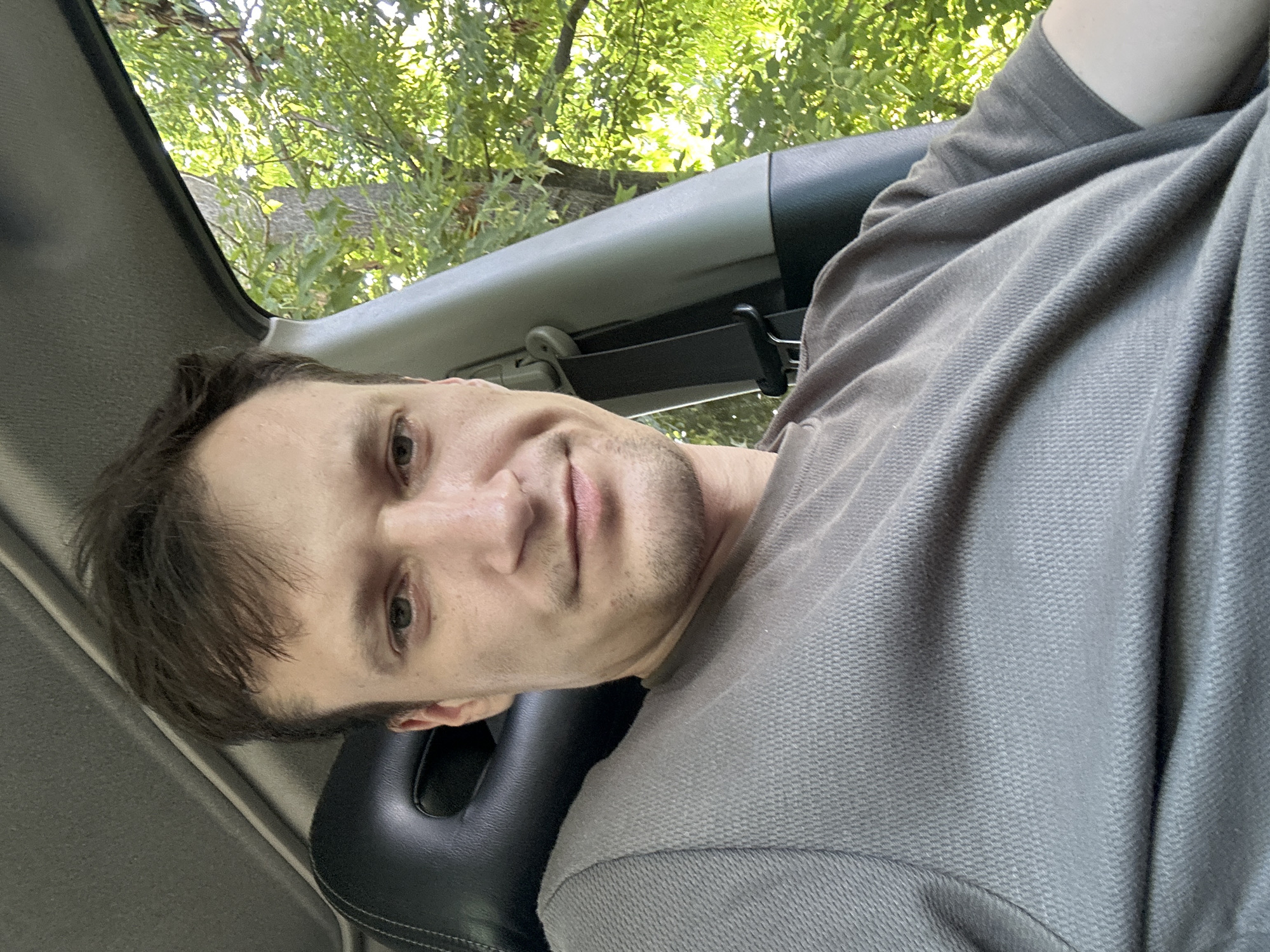A young man with short, dark brown hair and light skin is sitting in the driver's seat of a car, looking at the camera. He is wearing a grey t-shirt. The car interior, including a black headrest and grey trim, is visible. Sunlight filters through green trees outside the car window, suggesting an outdoor setting, possibly in Makiv, Ukraine, on a clear day.