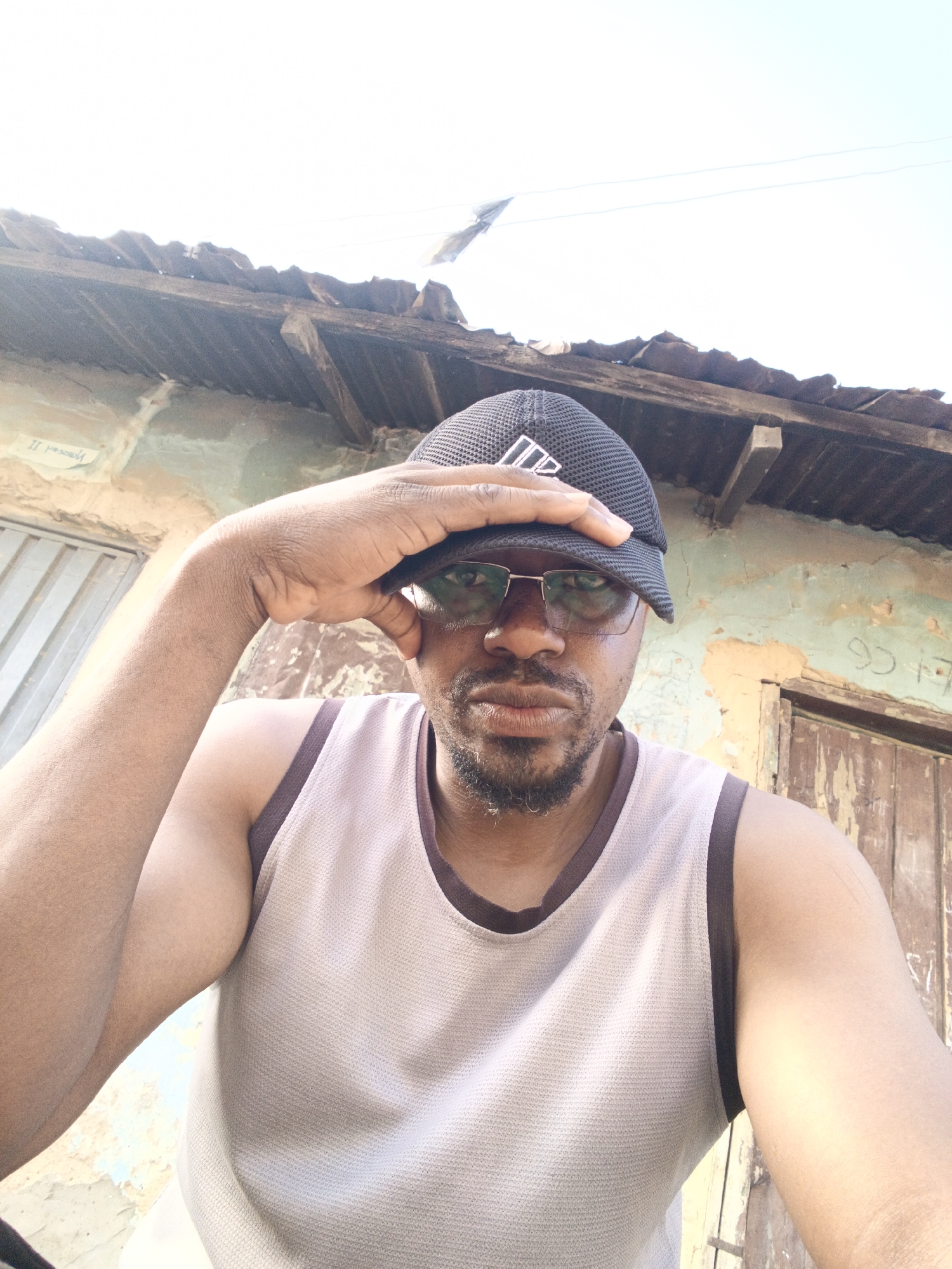A man stands outdoors in Gwagwarwa, Nigeria. He wears a sleeveless top, glasses, and a baseball cap, holding his hand up to his forehead. Behind him, a dilapidated building with a corrugated metal roof and peeling paint is visible. A weathered wooden door is also present.
