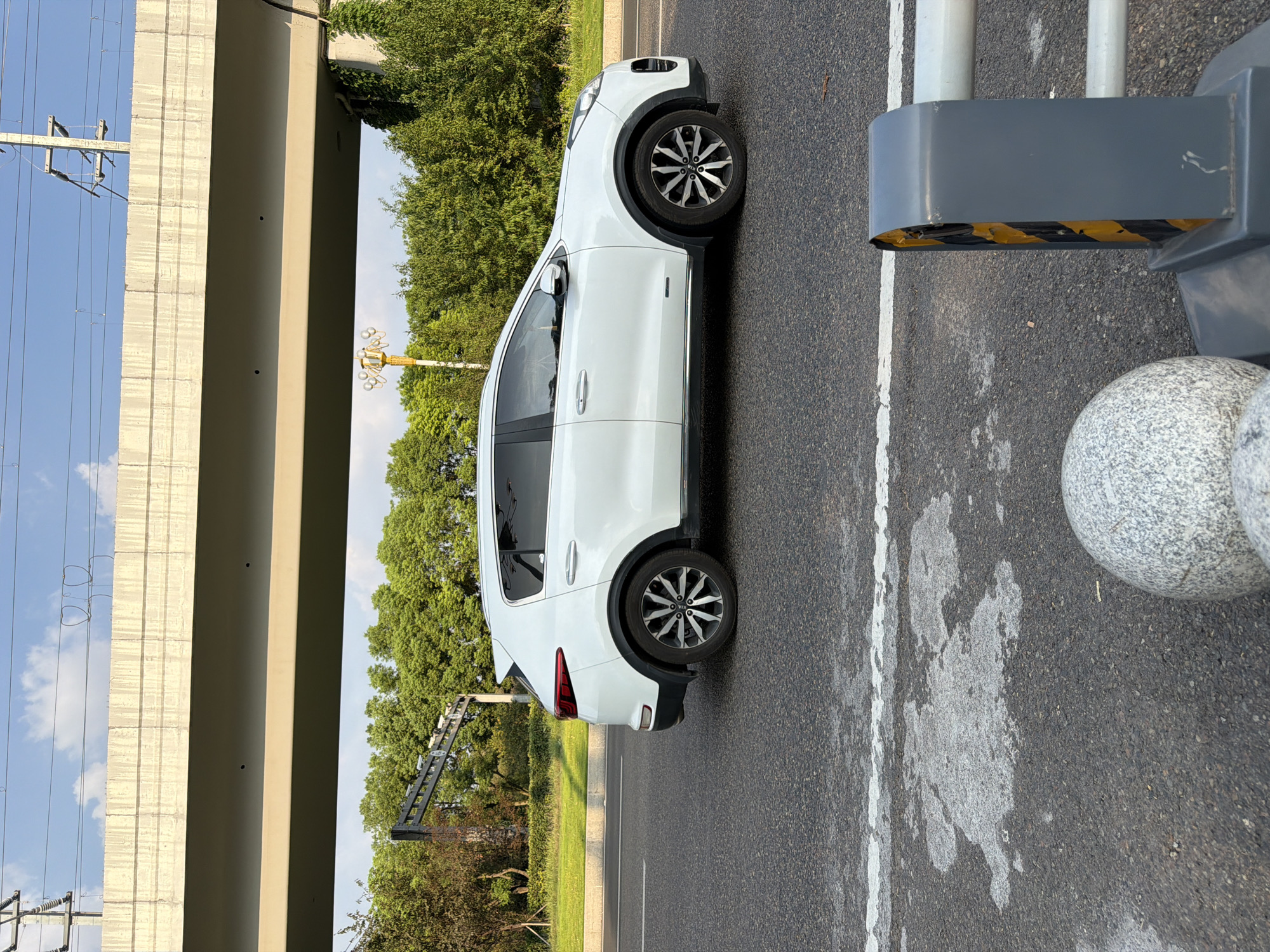 A white Kia Sportage is traveling on a road in Jiangjin, China, under an overpass. The car is viewed from the side, moving from left to right. The overpass is made of concrete and has overhead wires and utility poles visible above it. Lush green trees line the side of the road. In the foreground, a section of the road has a white line marking and what appears to be wet or stained asphalt. A gray and yellow traffic barrier is visible on the right side of the frame, along with a rounded, speckled boulder. The sky is blue with scattered clouds, and the sun is casting light, suggesting it is daytime.