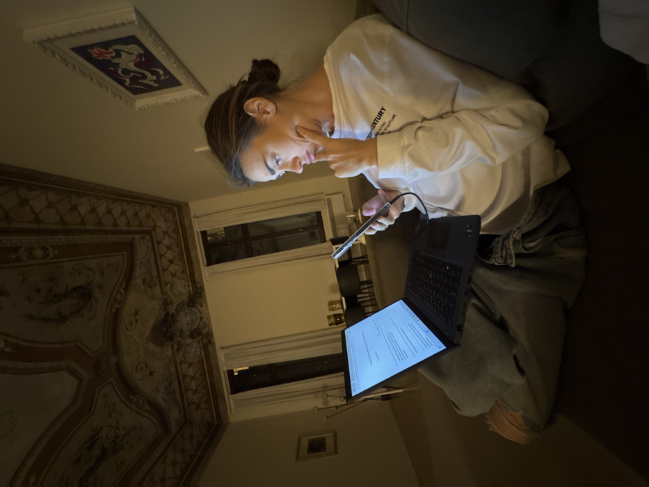 A female subject, with brown hair tied back, is seated on a dark sofa in a dimly lit room within a historic building in Venice, Italy. She is wearing a white long-sleeved shirt with text "21ST CENTURY SCULPTURE" and light-colored jeans. A black laptop is open on her lap, displaying an illuminated screen with visible text content. She holds an illuminated smartphone in her left hand, connected by a cable. Her gaze is directed towards the smartphone, while her right index and middle fingers are pressed against her right cheek, indicating a pensive or focused state. The room features a high, ornate ceiling adorned with frescoes. In the background, two tall windows with white curtains are visible, reflecting interior lights. Further back, a dining area with dark chairs and a table can be seen, along with a floor lamp and two framed artworks on the walls.