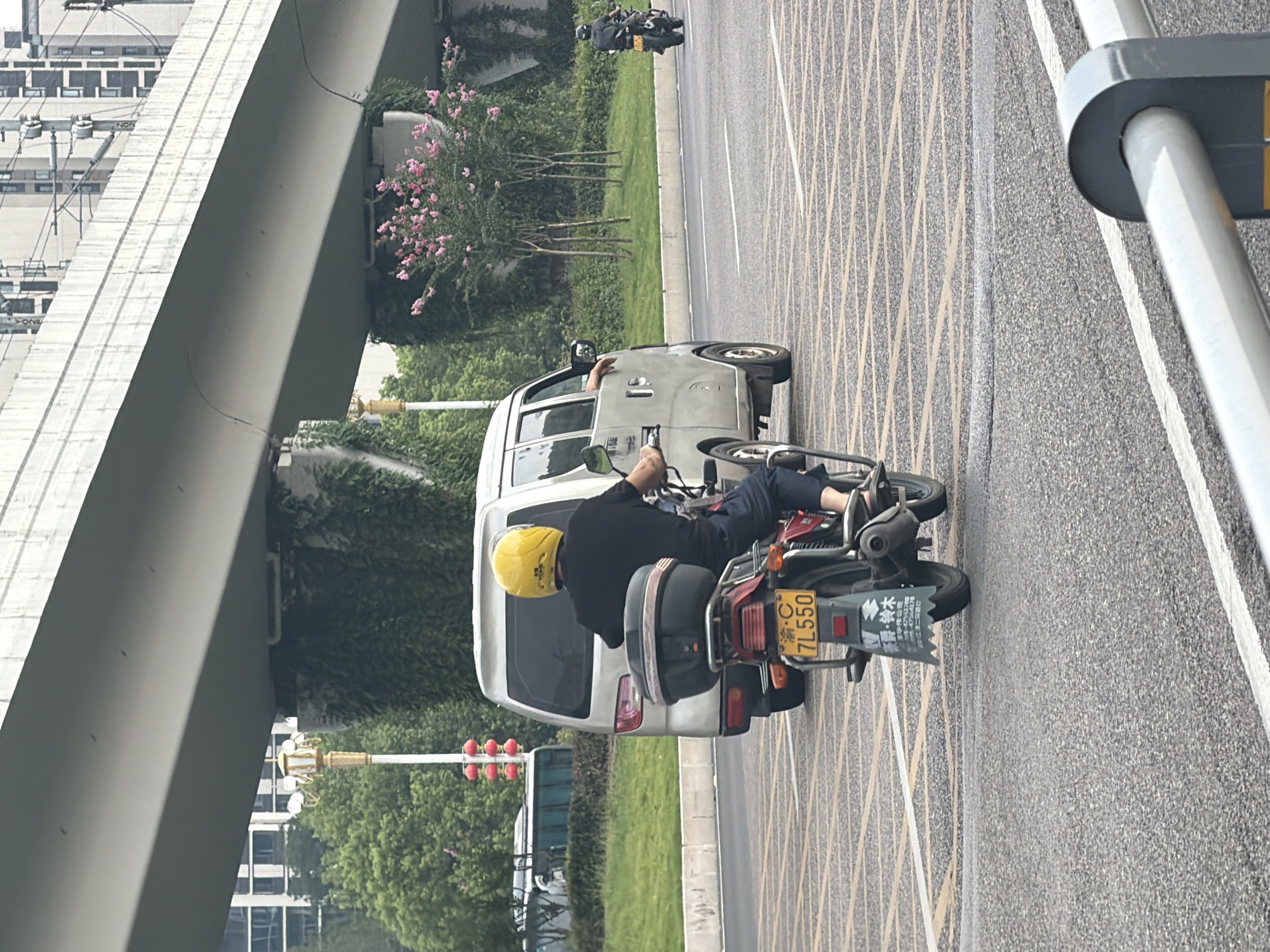 In Jiangjin, China, on a cloudy day, a person wearing a yellow helmet and black t-shirt rides a motorcycle. The motorcycle has a license plate that reads "渝C 7L550". Behind the motorcycle, a silver van is parked. The vehicles are on a multi-lane road with textured yellow markings. A modern elevated highway structure with concrete supports covered in ivy and flowering pink bushes looms overhead. In the background, a building with multiple windows and a bus are visible. Another motorcycle is seen in the distance on the right side of the road. A gray barrier is in the foreground.