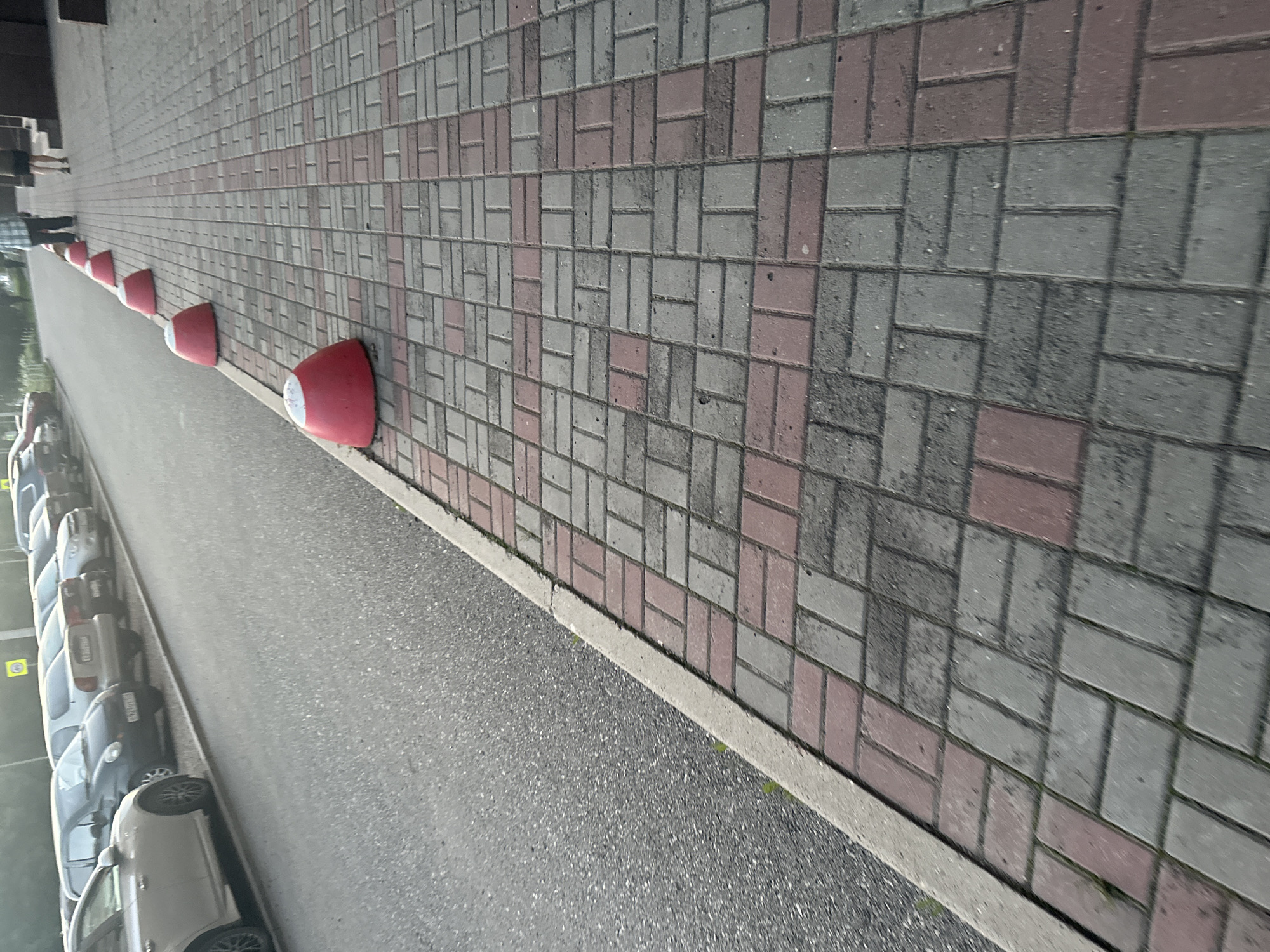 A row of red bollards with white tops are lined up on a patterned brick walkway. The walkway is made of rectangular bricks in alternating shades of grey and pink. To the left of the walkway is a paved area with a row of parked cars. The cars are of various colors including white, grey, and black. The scene appears to be outdoors, likely a street or parking lot. The lighting suggests it is daytime. There is a slight tilt to the image, making it appear as if it was taken from a slightly elevated or angled perspective. No text is clearly visible in the image.