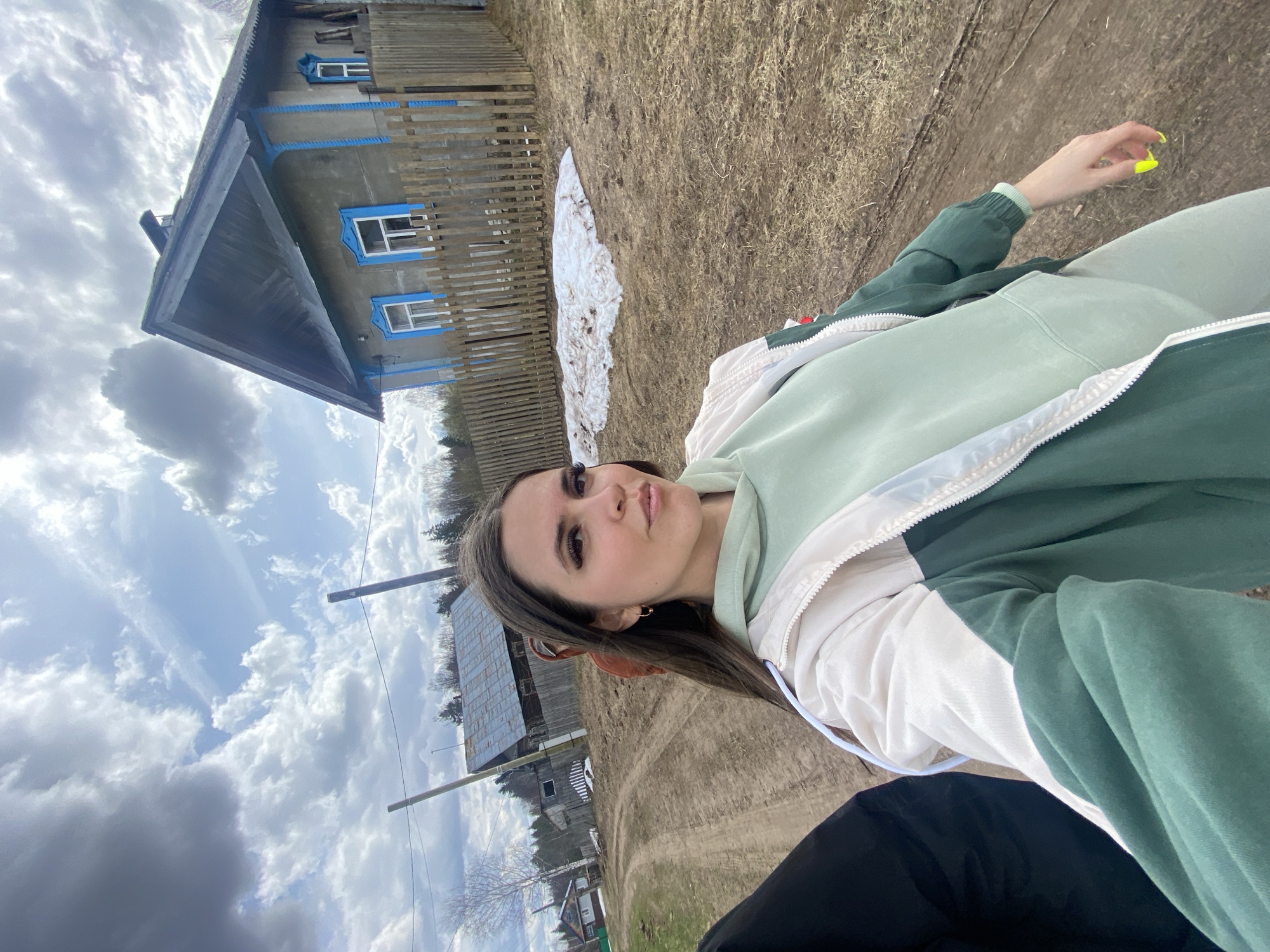 A woman with brown hair and brown eyes is positioned in the foreground of a horizontally rotated image. She is wearing a green and white tracksuit. Her left hand is extended with neon yellow nail polish visible. In the midground, there is a weathered wooden fence and a dilapidated gray wooden house with blue window frames. Patches of snow are visible near the house. In the background, a cloudy sky dominates the upper half of the image. Below, additional houses and trees can be seen, indicating a rural setting. The environment suggests the location is Zhigalki, Russia.