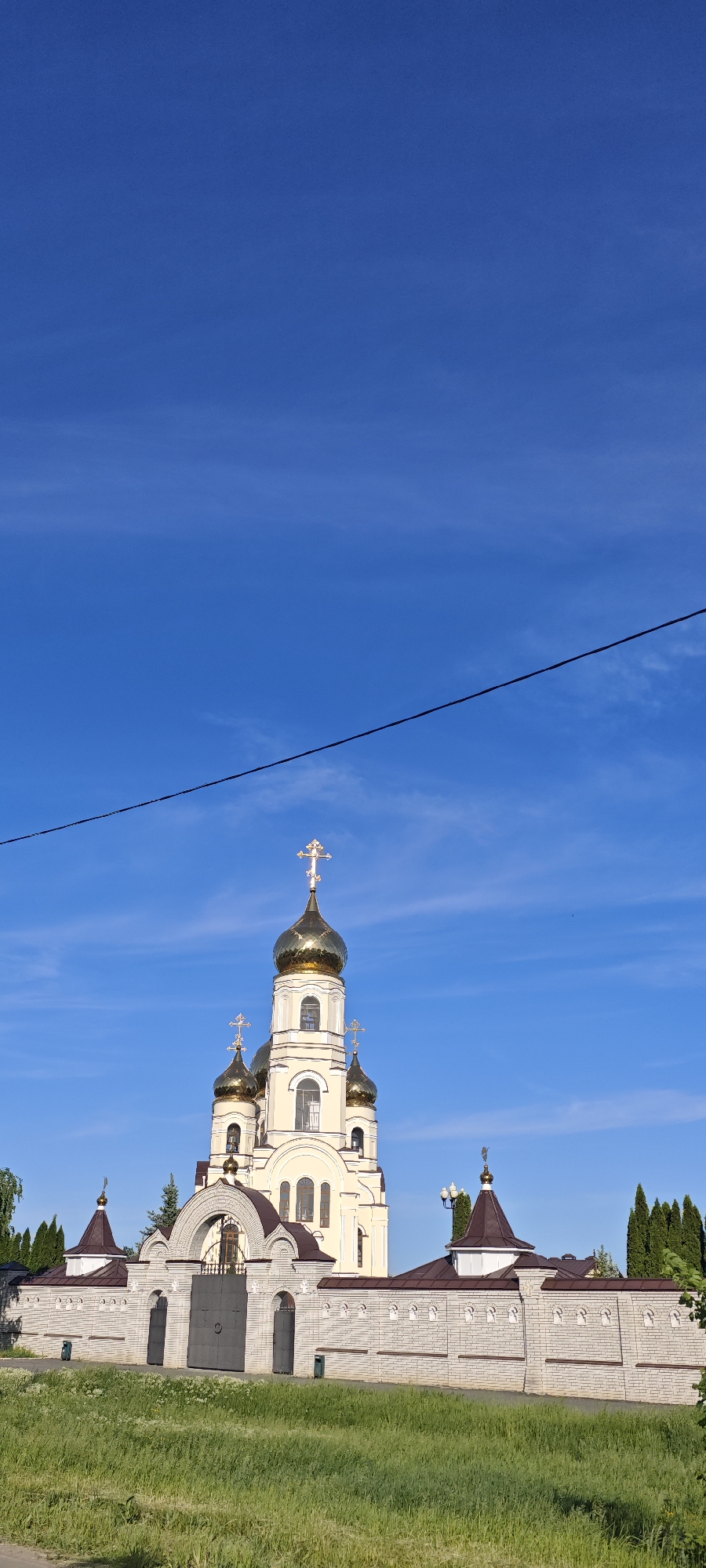 A vertical, eye-level shot depicts a church complex situated in Alekseyevka, Russia. The main structure is a cream-colored church with multiple golden domes, each topped with a cross. A brick wall, punctuated by arched gateways and topped with brown roofs, surrounds the church. A black metal gate is visible in one of the gateways. The foreground is a grassy area. Overhead, a clear blue sky dominates the upper portion of the frame, with a single black power line cutting diagonally across it.
