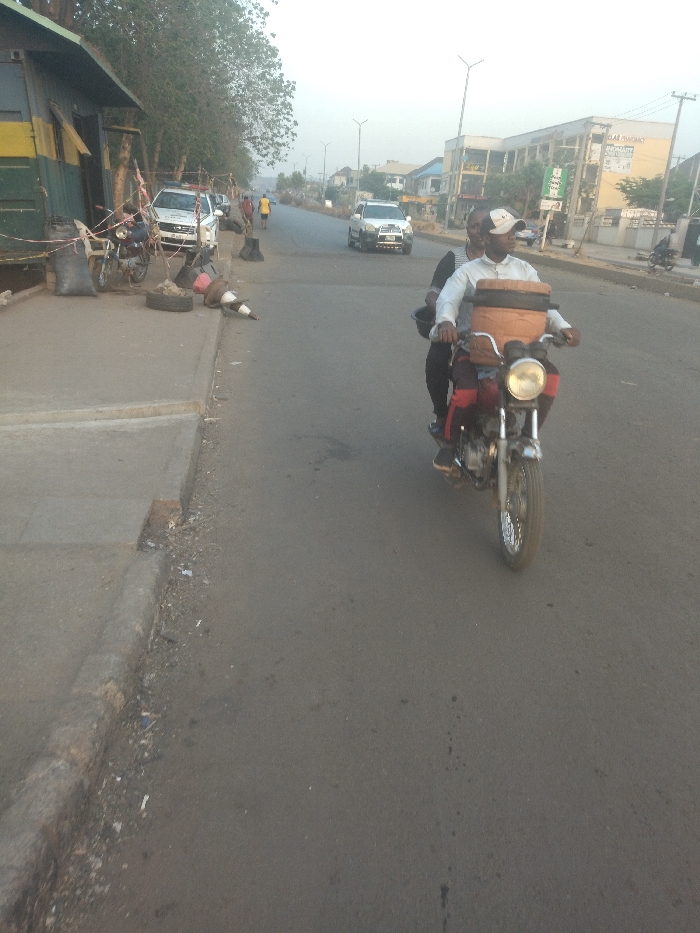 The image shows a street scene in New Karu, Nigeria (latitude: 8.998, longitude: 7.571). There is a man driving a motorcycle with another person riding as a passenger. The motorcycle has a large, brown container strapped to the back. The motorcycle is driving on a paved road in the middle of the image, heading towards the right side of the image. There are buildings on the right side of the road and trees on the left. The scene is taken during the day. There is a parked car on the left side of the road, near a sidewalk. There is a building on the left side of the image. There are traffic cones and a tire on the sidewalk. There are two people walking on the sidewalk on the left side of the image. The sky is a light gray. The ground is covered in asphalt and there are patches of dirt. The image appears to be taken from a distance and angled slightly downwards.