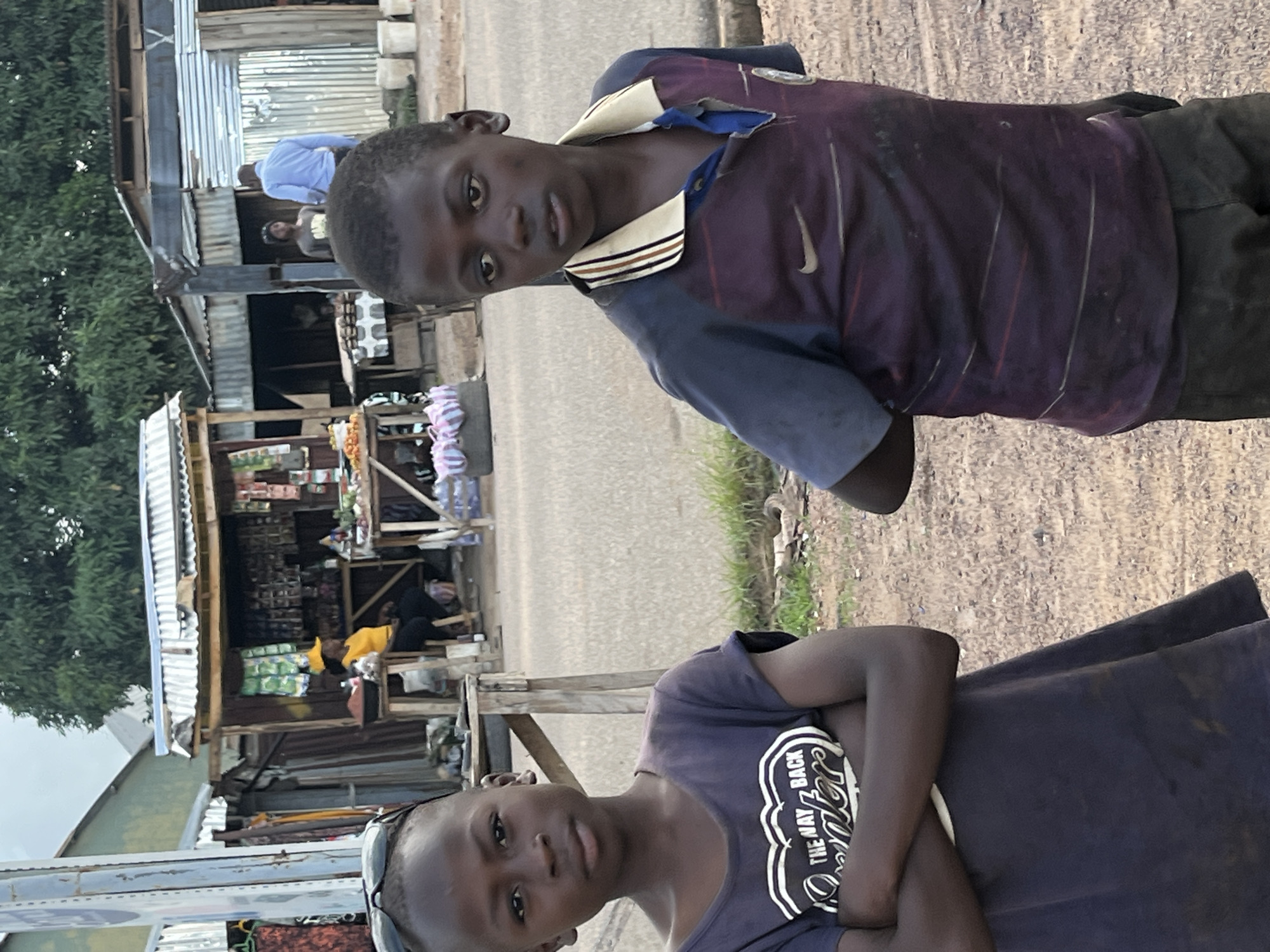 The image depicts a street scene in Barakin Akawo, Nigeria. Two young boys are in the foreground. The boy on the left, facing forward, wears a dark t-shirt with a white graphic and has his arms crossed. The boy on the right is looking slightly to his right, wearing a purple and blue collared t-shirt with a Nike logo. Behind them, a roadside market is visible, with stalls selling various goods. Several people are present at the market, including a woman in a yellow shirt and hat sitting at a stall, and two men, one in a blue shirt and another in the background. The environment appears to be dusty with some vegetation and makeshift structures.