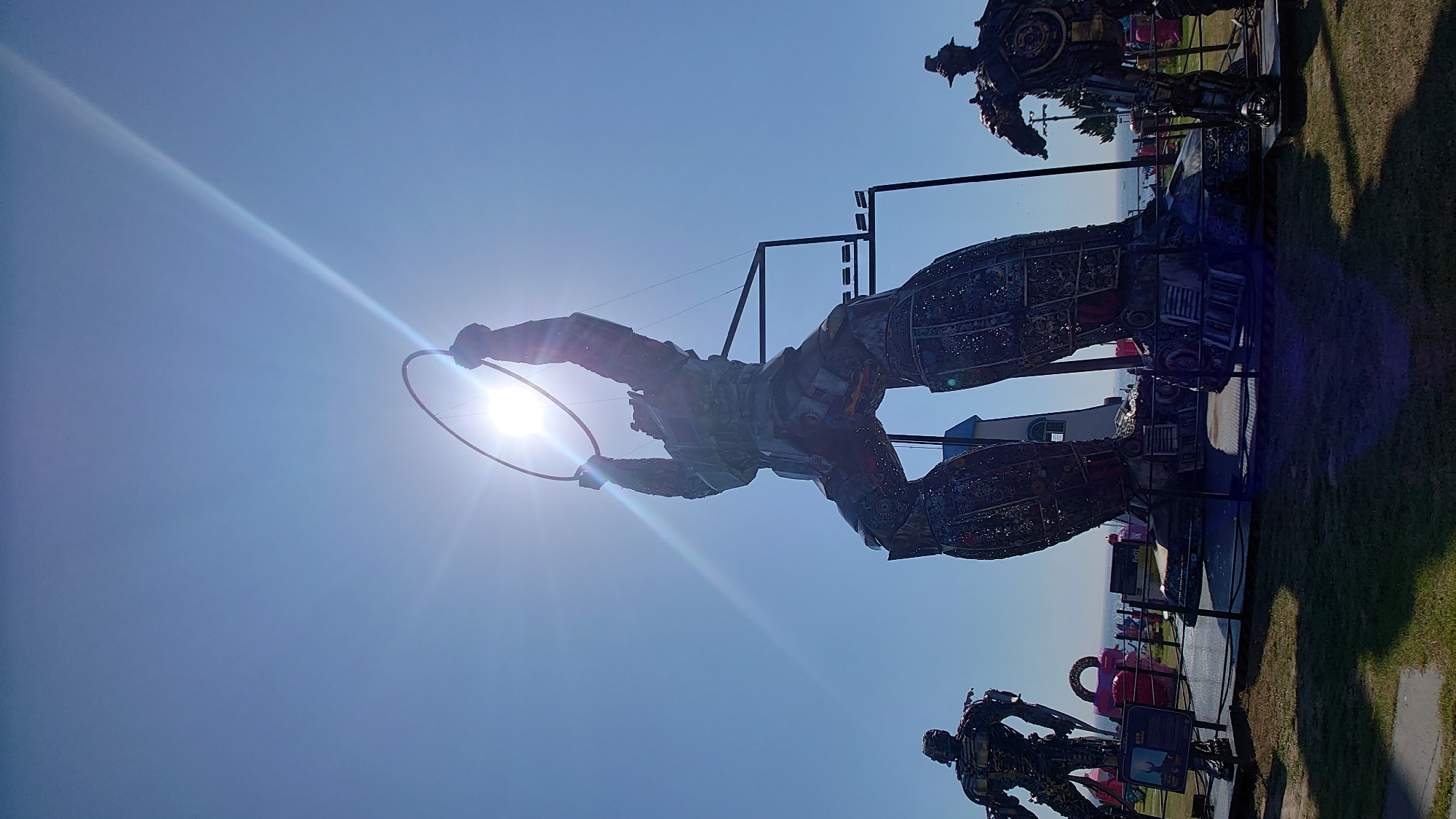 The image captures a bright, sunny day with a clear blue sky in Ulsan, South Korea. The sun is high and creates lens flare effects. The main focus is on a large, imposing sculpture of a robot-like figure made from metal parts, standing with its arms raised, holding a circular object, possibly a ring or a hoop, against the sun. The sculpture is detailed and appears to be constructed from recycled materials.

To the left of the central sculpture, another similar, but smaller, robot sculpture is visible, also standing on a platform. To the right, a third robot sculpture is partially visible. The sculptures are placed on a grassy area with some paved pathways. In the background, beyond the sculptures, there is a glimpse of the sea, suggesting the location is coastal. There are also some pink structures and a lighthouse-like building visible behind the central sculpture. The overall scene depicts an outdoor art installation, likely in a park or public space in Ulsan.