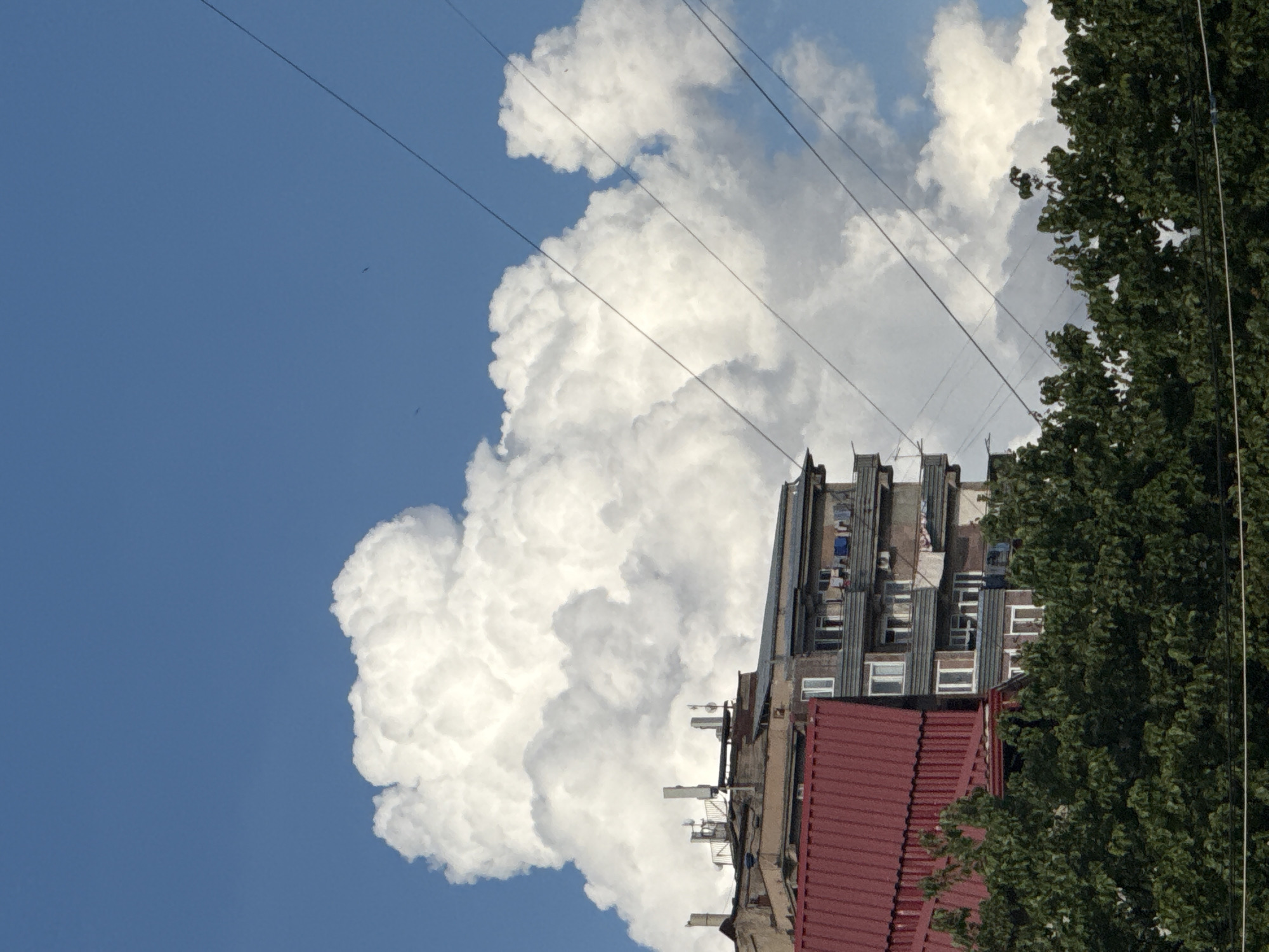 The image depicts a cloudy sky above a multi-story building in Abovyan, Armenia. The building has a red-tiled roof in the foreground, with an adjacent concrete structure featuring multiple balconies with laundry hanging. Several antennas and communication towers are visible on the rooftop of the building. Dark green trees line the bottom of the frame, partially obscuring the lower levels of the buildings. Several power lines stretch diagonally across the sky. The sky is a bright blue, with large, white cumulus clouds dominating the upper portion of the image. There are no people visible in the scene.